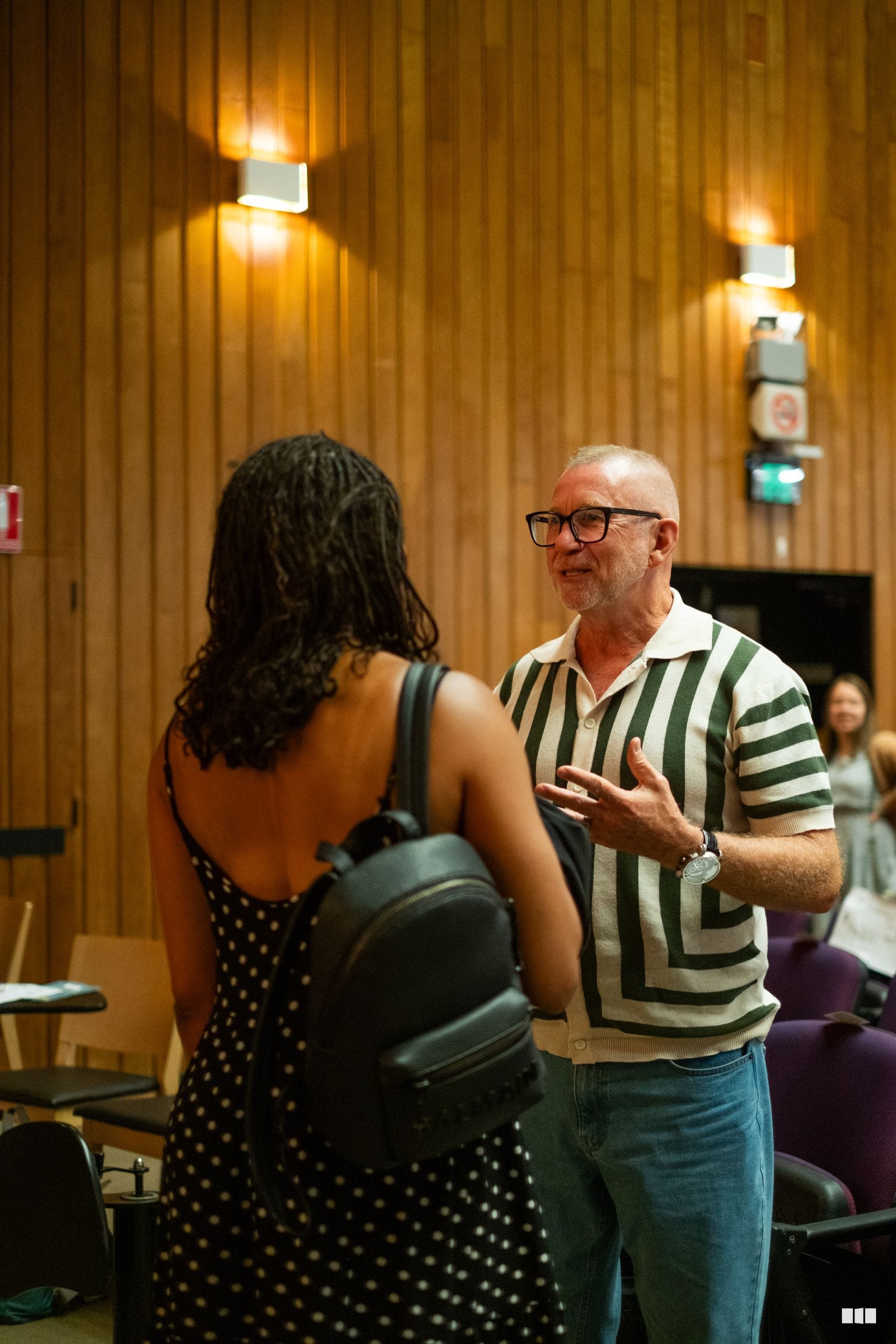 A man in a green and white striped shirt engages in conversation with a woman at an indoor venue with wood-paneled walls.