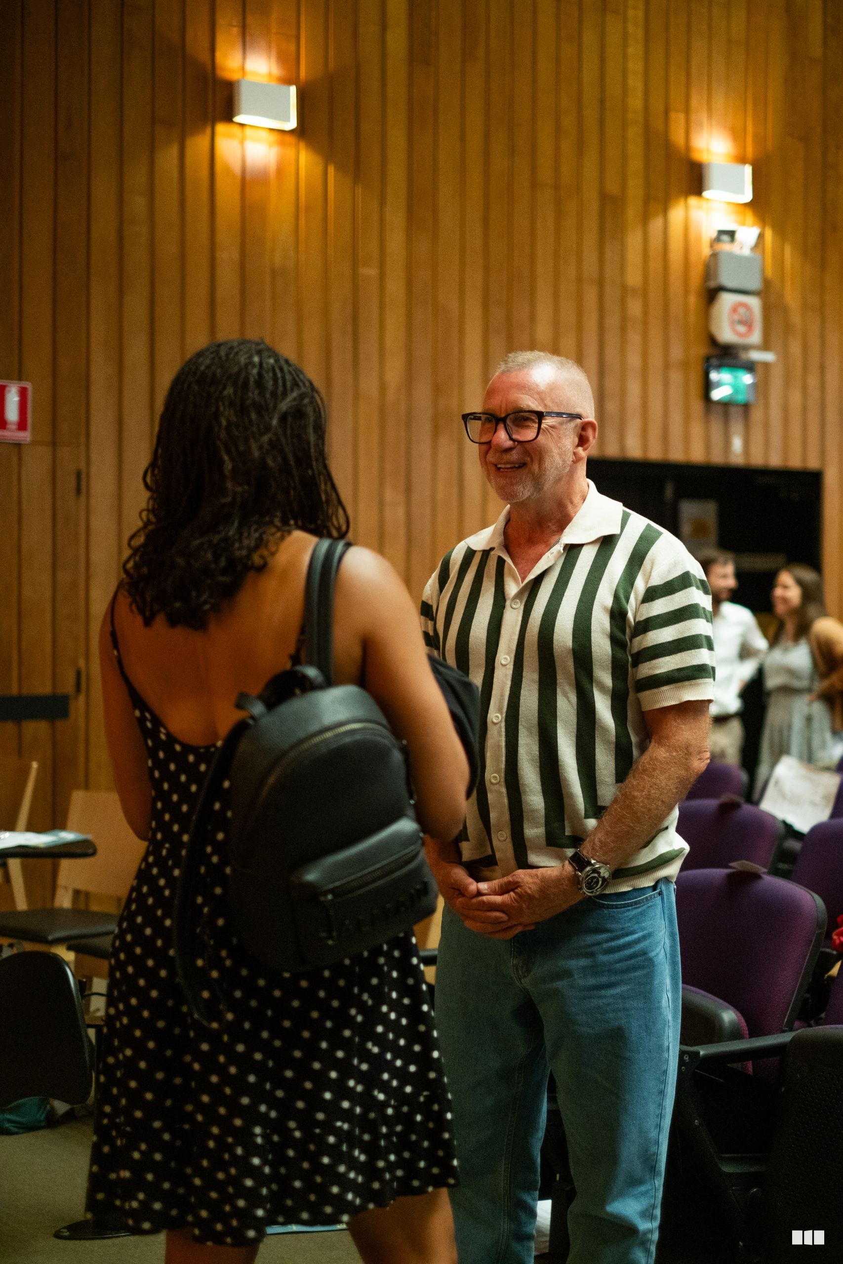 A man in a green and white striped shirt converses with a woman at an indoor event in a wood-paneled venue.