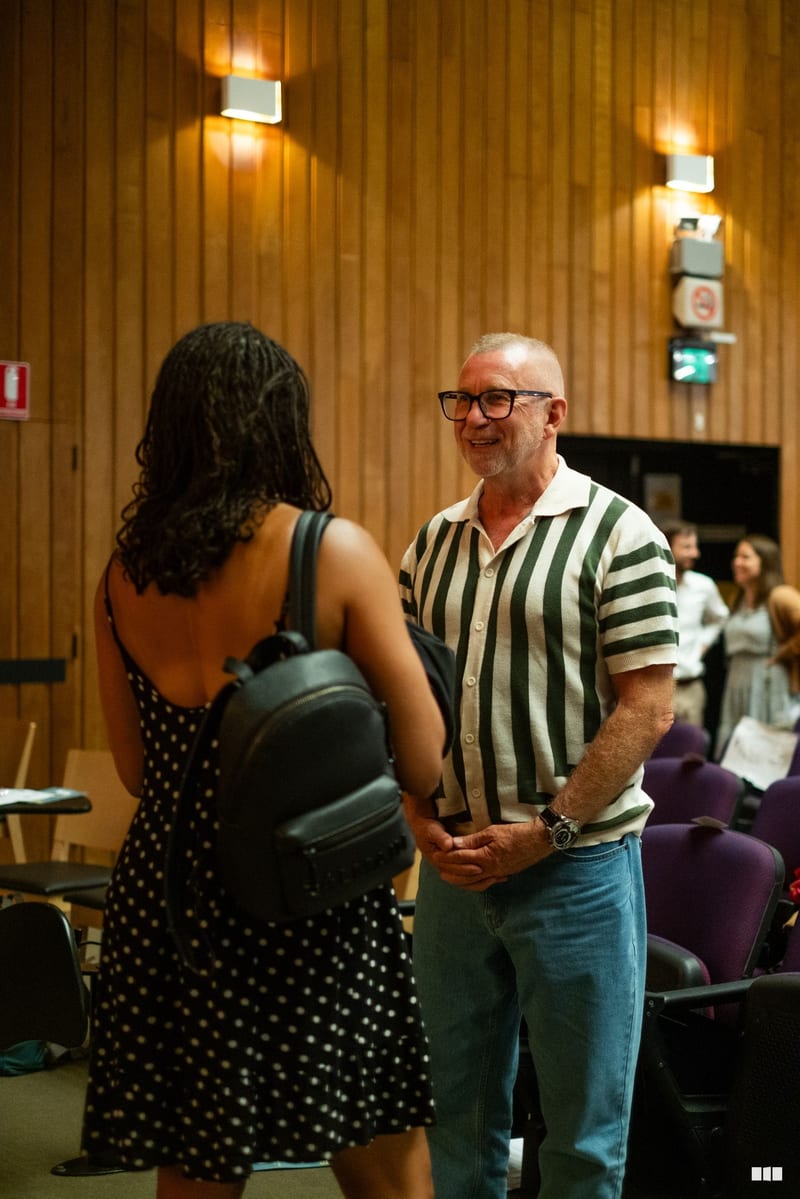 A man in a green and white striped shirt converses with a woman at an indoor event in a wood-paneled venue.