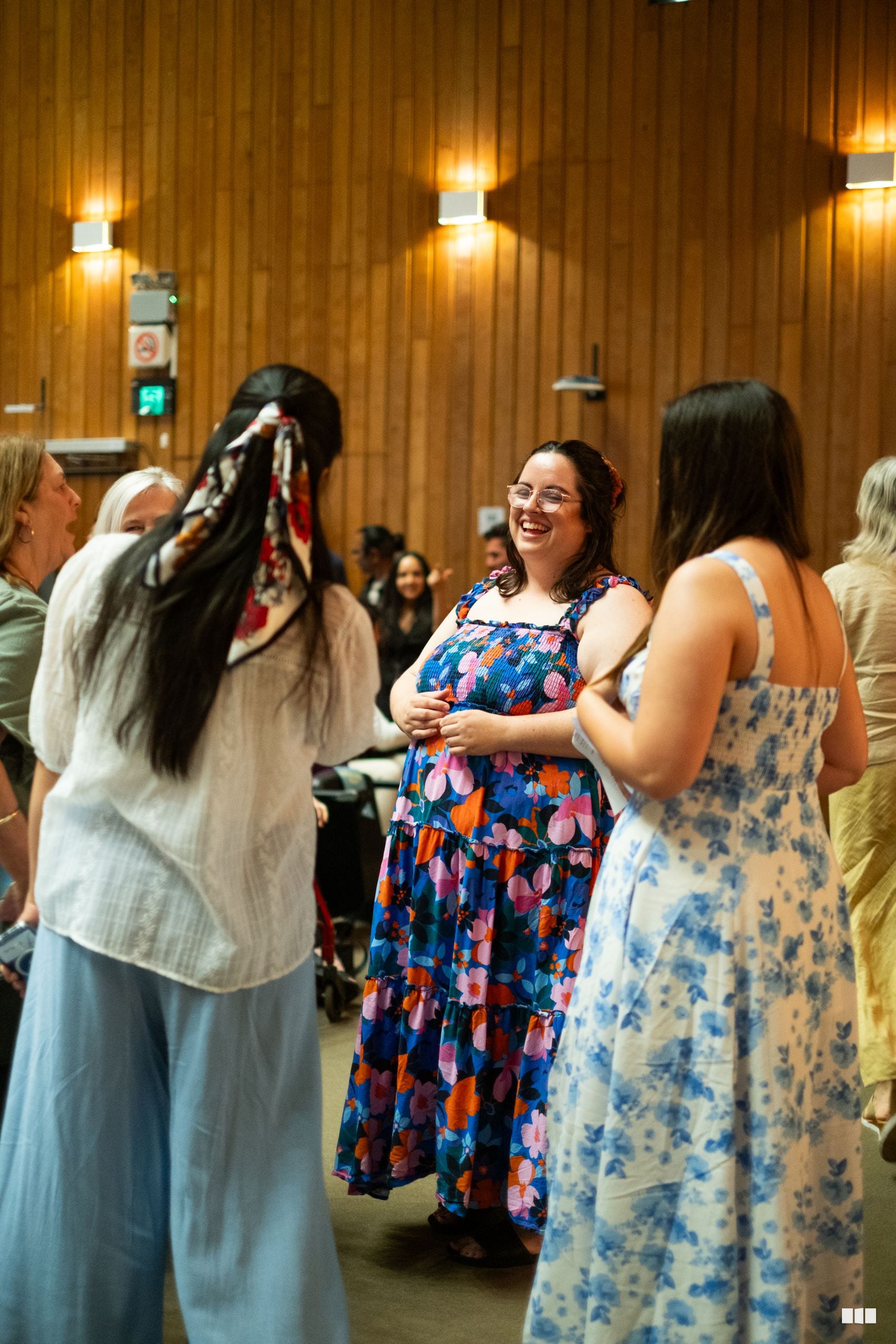 A woman in a colorful floral dress smiles while conversing with others in a wooden-paneled venue during an informal social gathering.