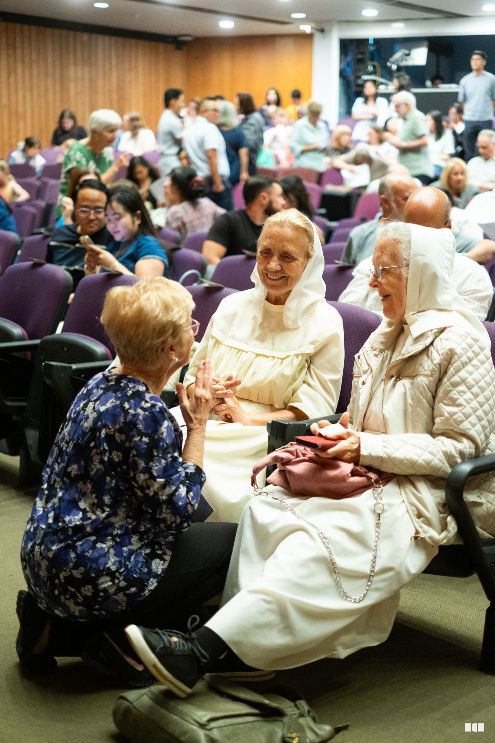 Three elderly women in white head coverings share a warm conversation and smile together at a community gathering in an auditorium.