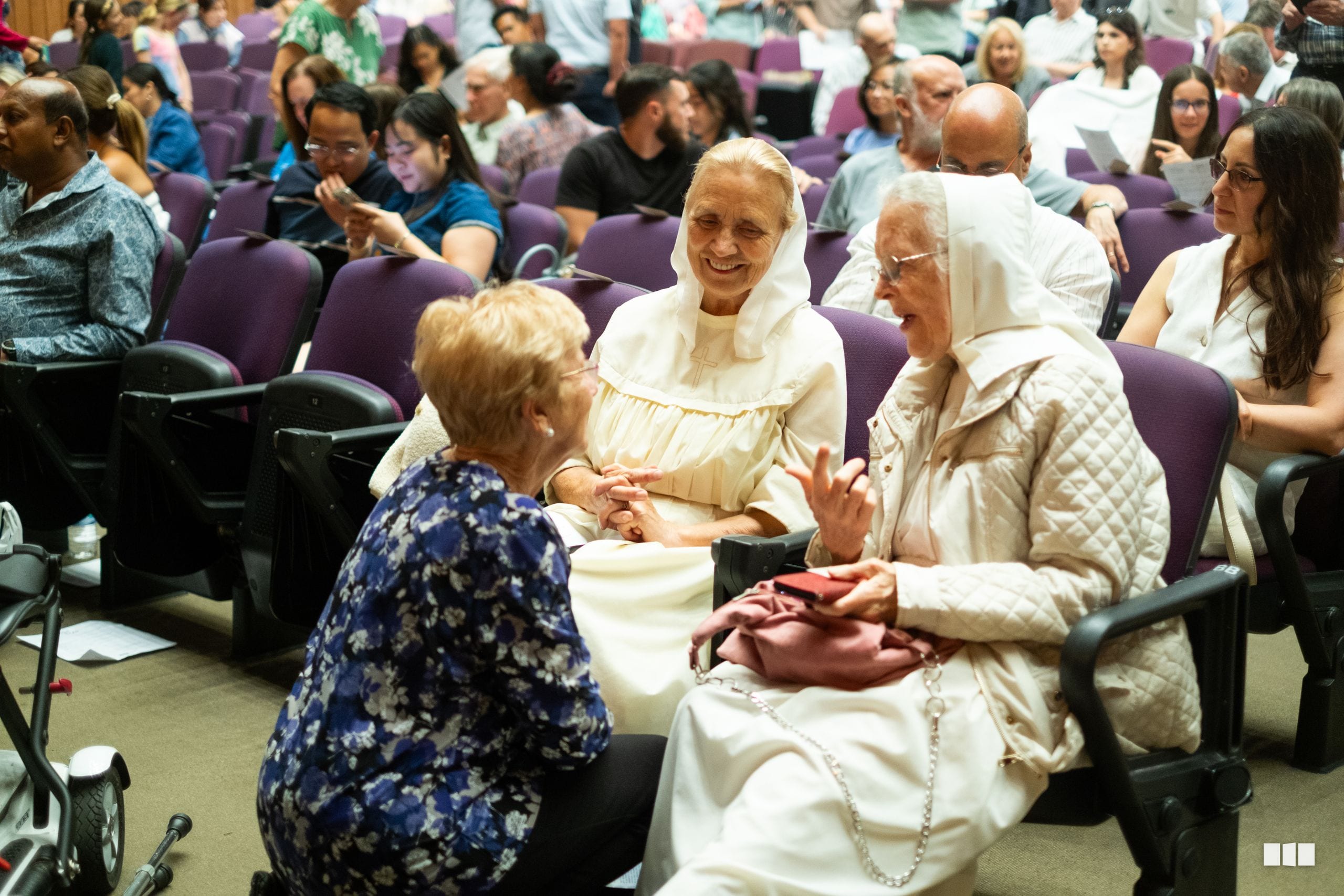 Three elderly women in white ceremonial dress share a joyful moment of connection with a seated attendee in an auditorium setting.