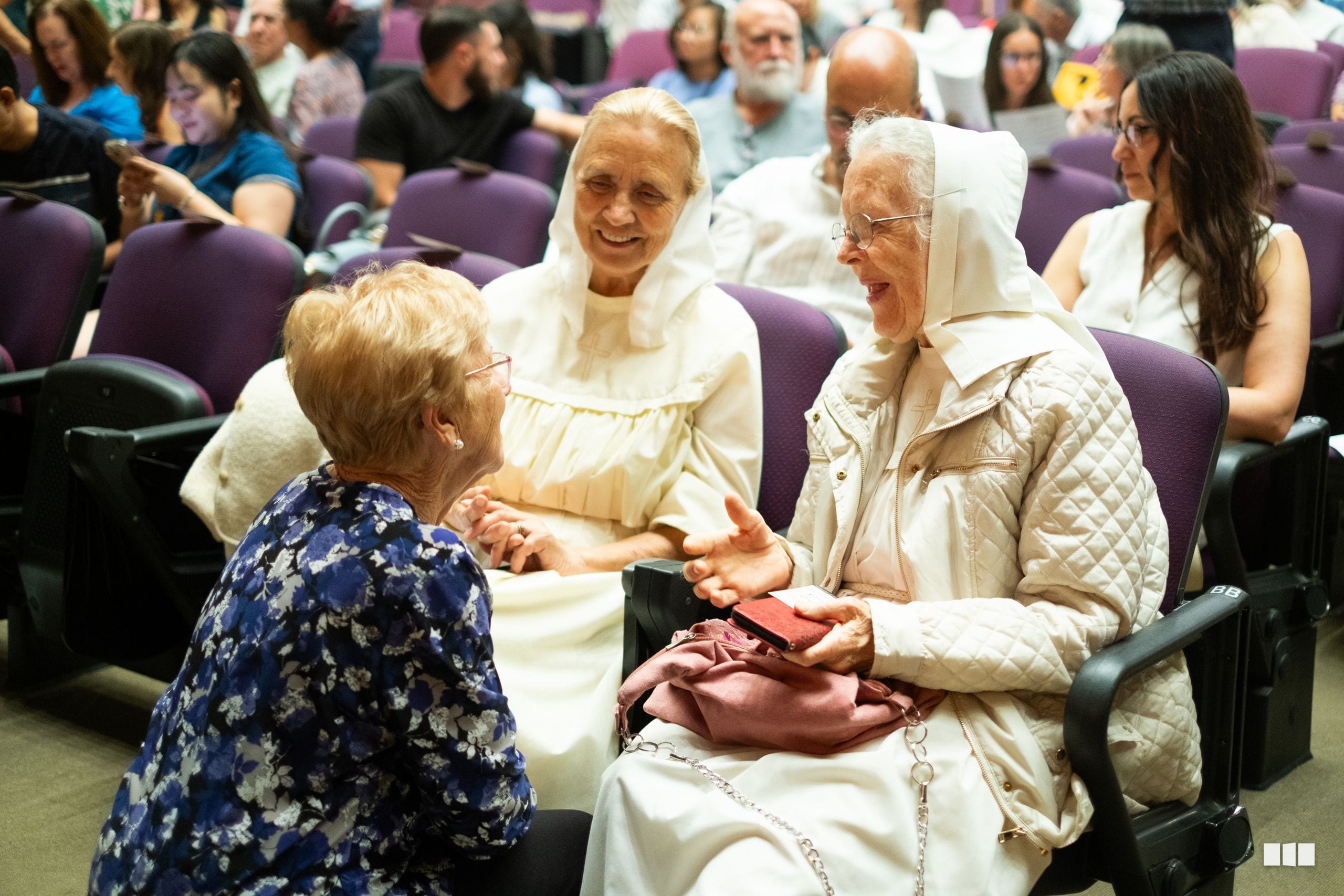 Elderly women in religious attire engaged in warm conversation with an audience member at a community gathering.