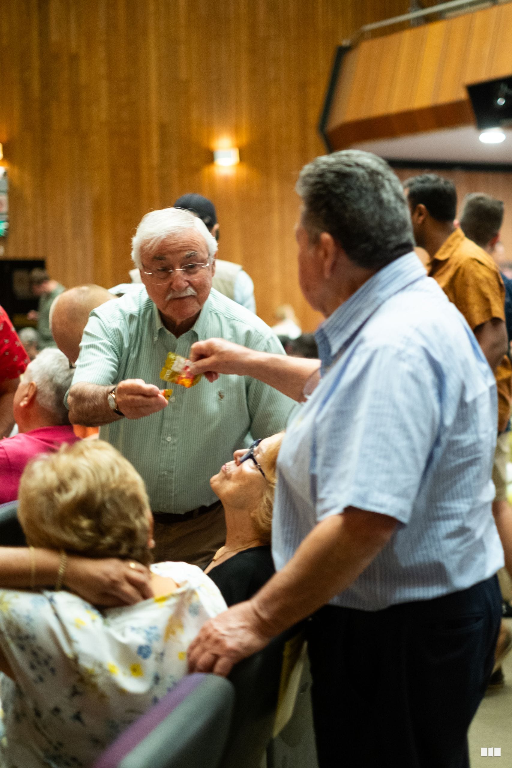 Elderly man in glasses shares food with seated guests at an indoor community gathering in a wooden-paneled venue.