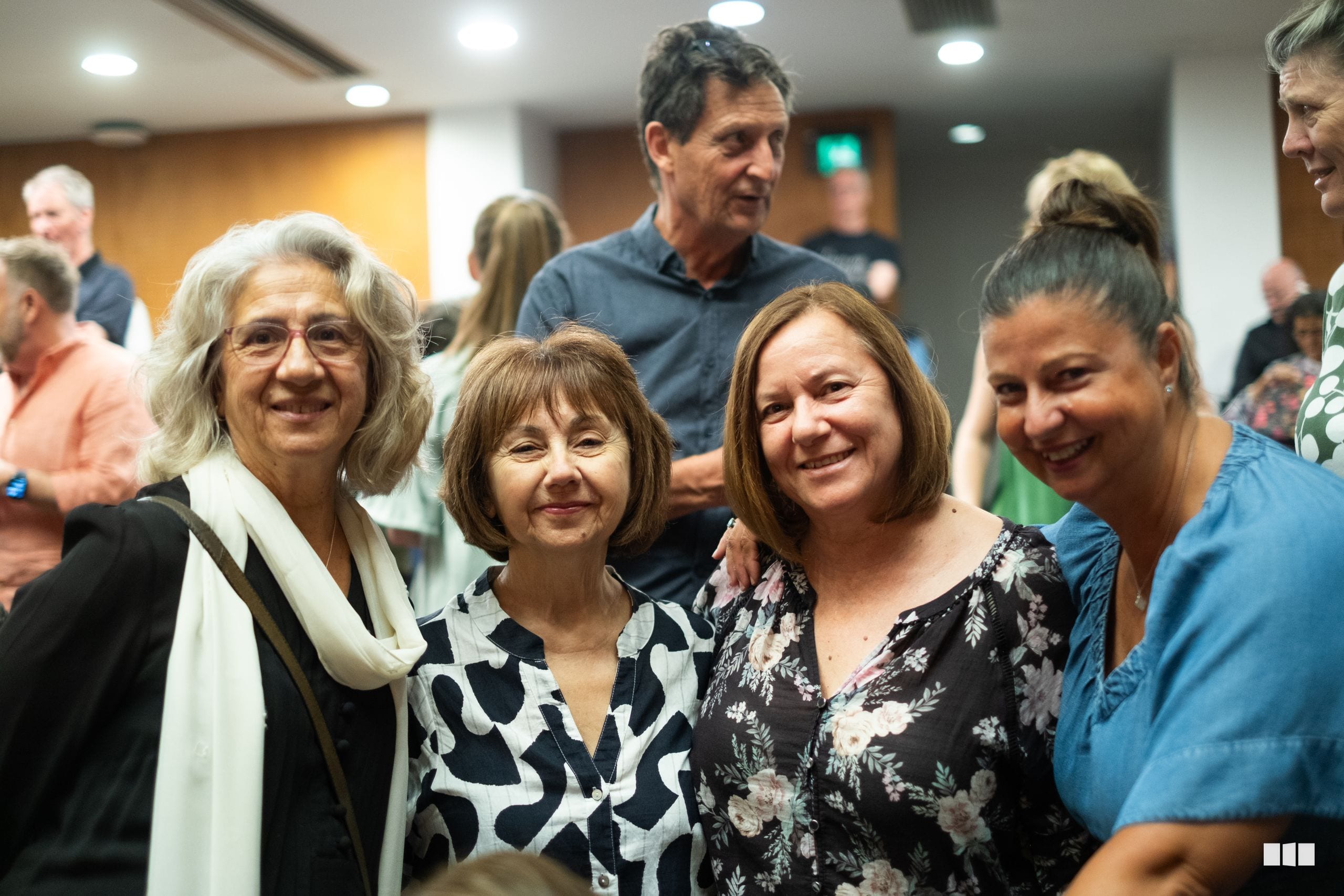 Four women smiling together at an indoor social gathering with other attendees visible in the background.