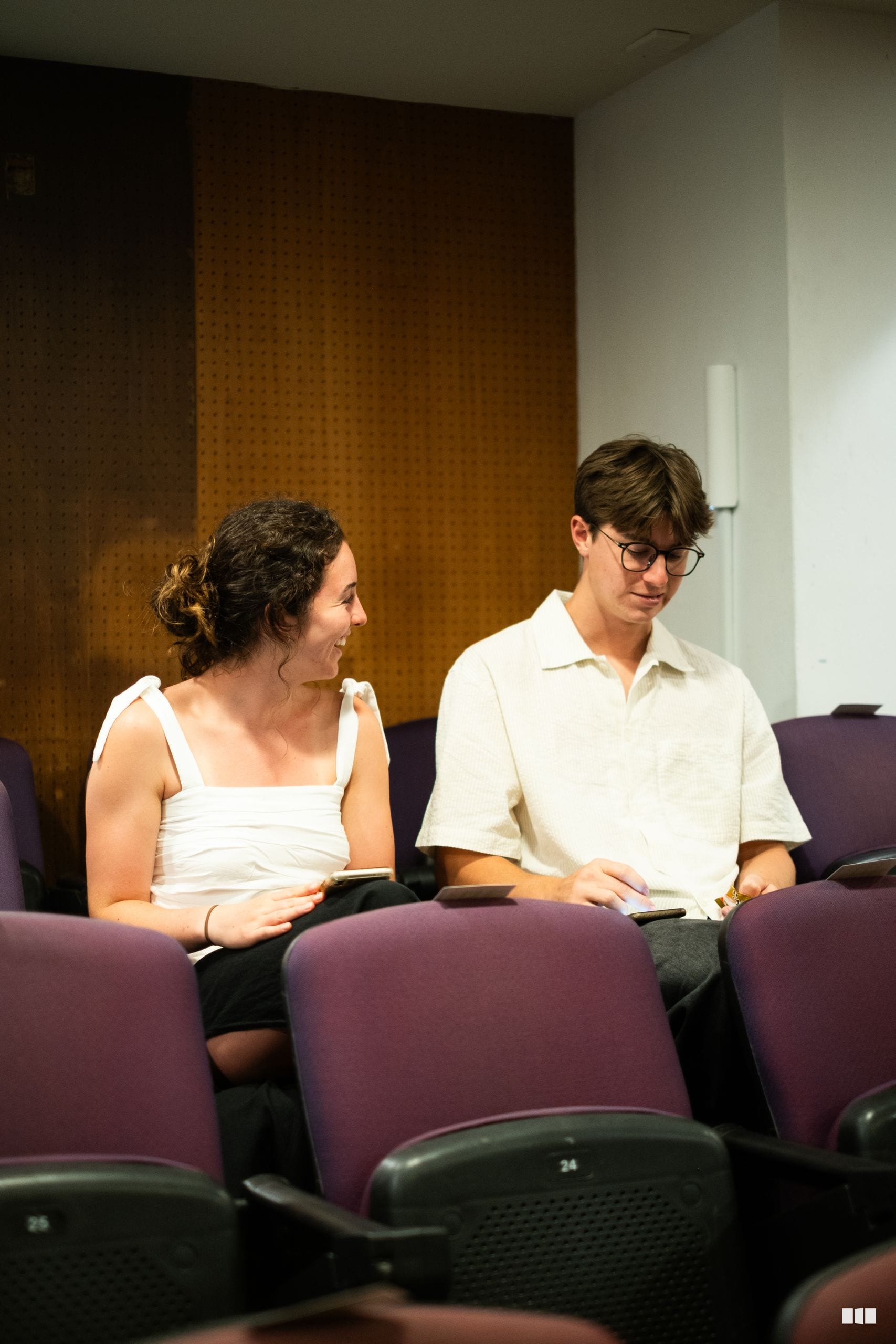 Two young adults sitting together in an auditorium, each holding mobile devices and smiling during what appears to be a rehearsal or event.