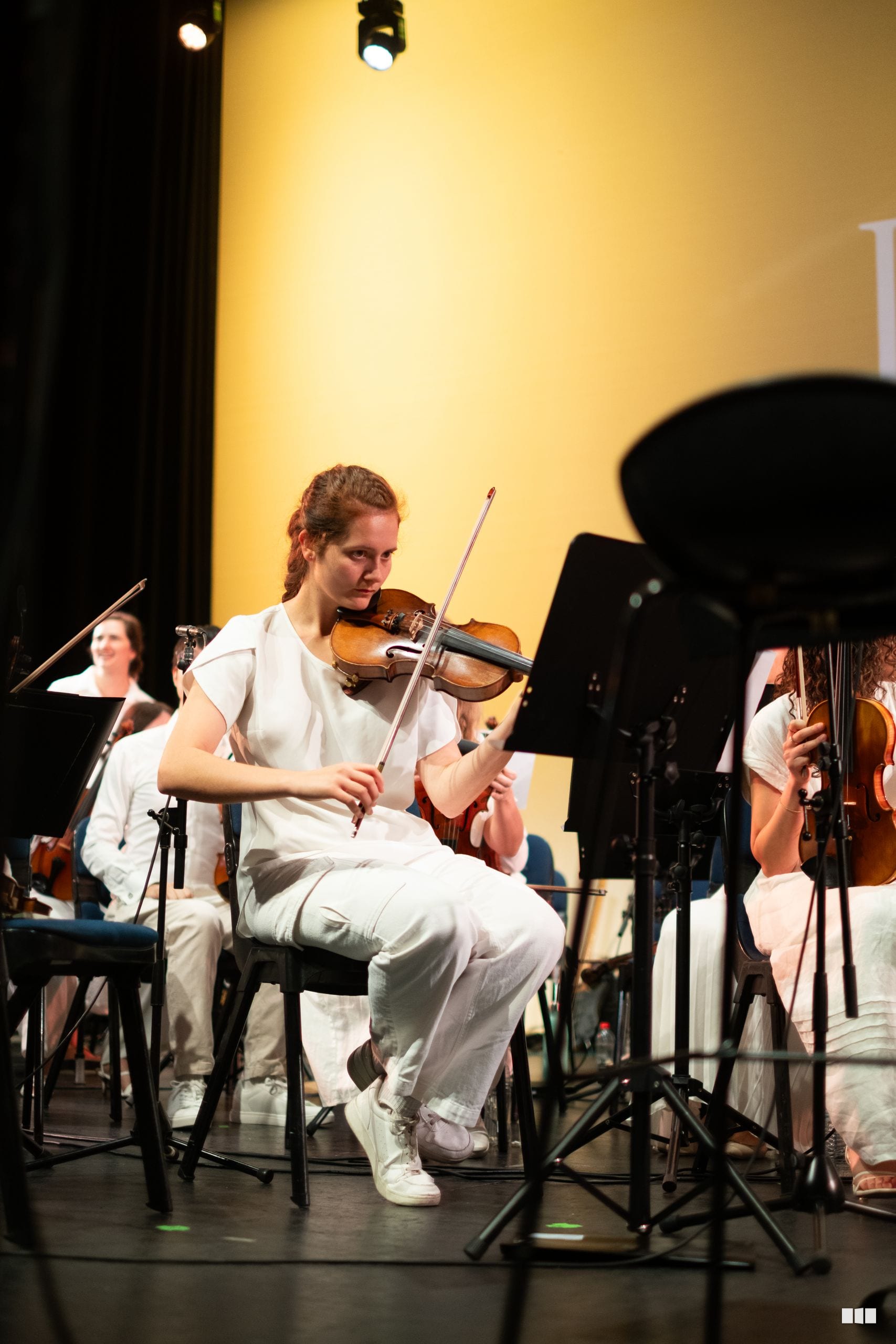 A young violinist performs on stage during an orchestral concert, focused on her instrument with other orchestra members visible in the background.