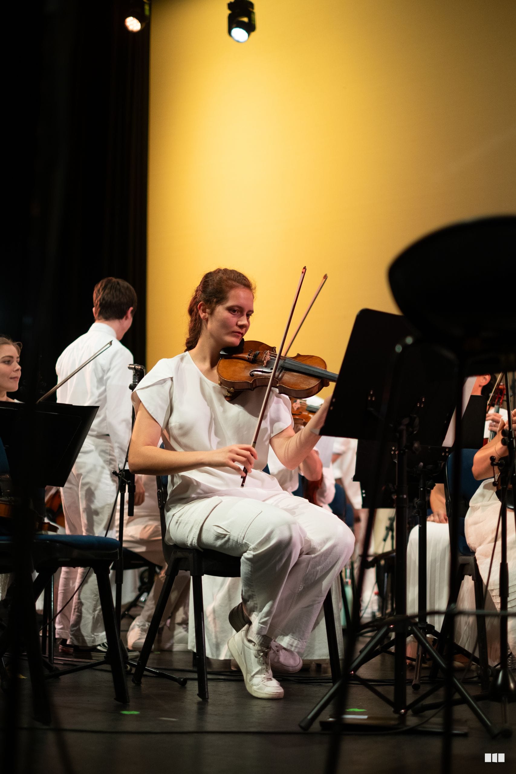 A young violinist performs during an orchestral concert, seated among ensemble members against a warm yellow-lit backdrop.