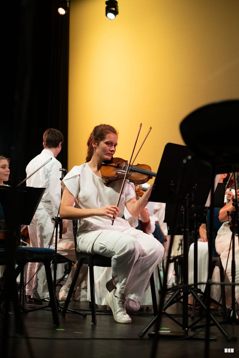A young violinist performs during an orchestral concert, seated among ensemble members against a warm yellow-lit backdrop.