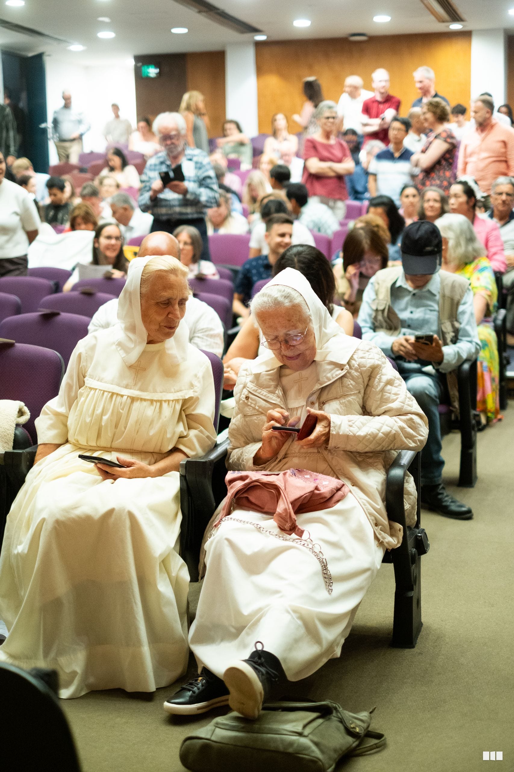 Two elderly women in traditional white religious dress sit together in the front row of a crowded community gathering or service.