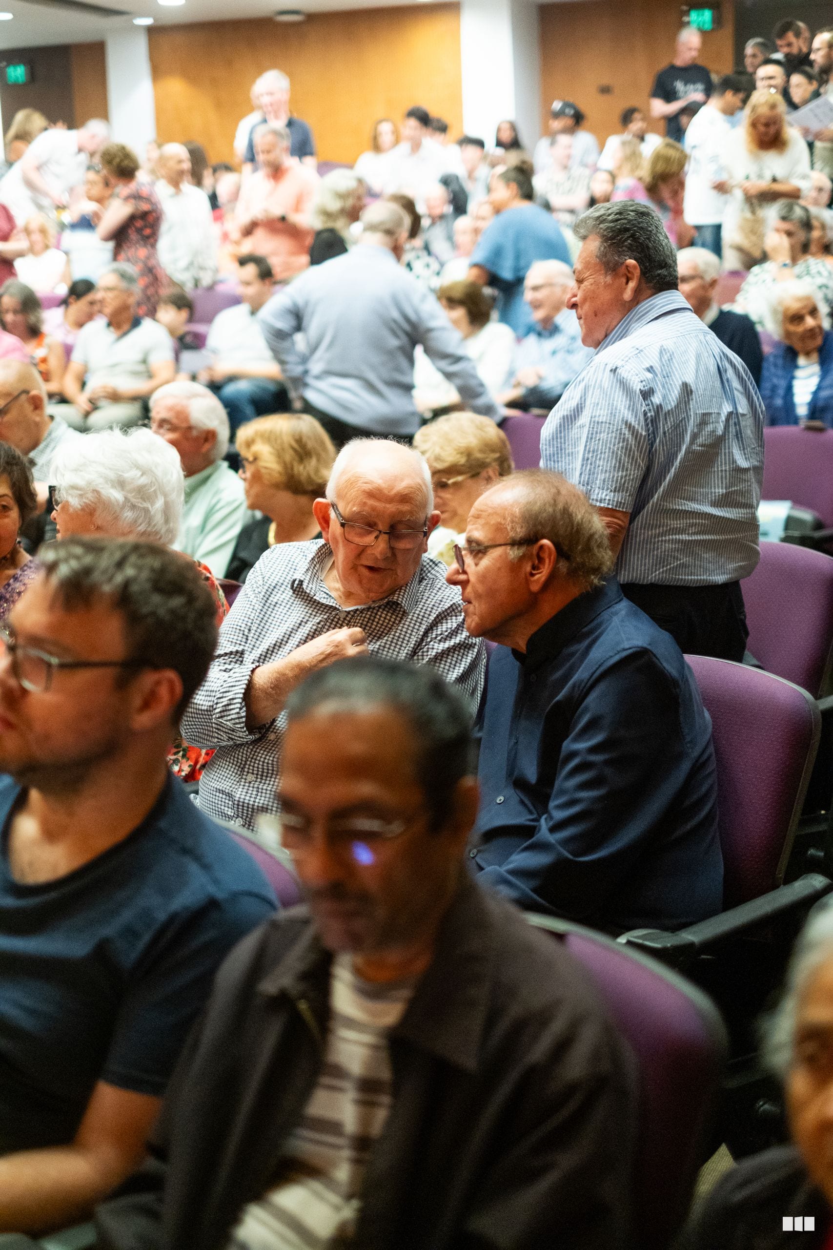 A diverse audience of mostly older adults sits together in a community venue during what appears to be a public gathering or performance event.