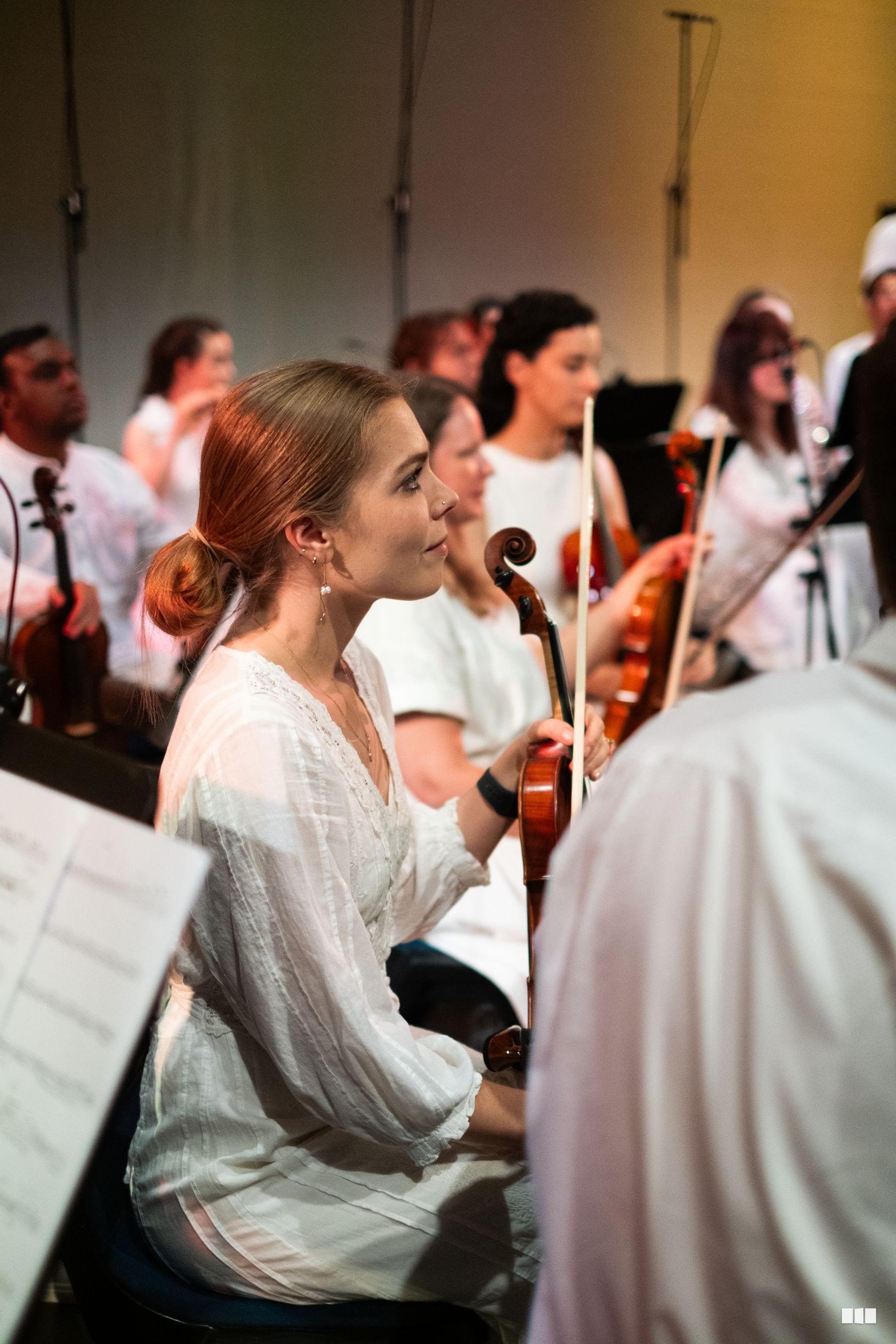 A violinist in white clothing performs with orchestra members during a rehearsal or concert.
