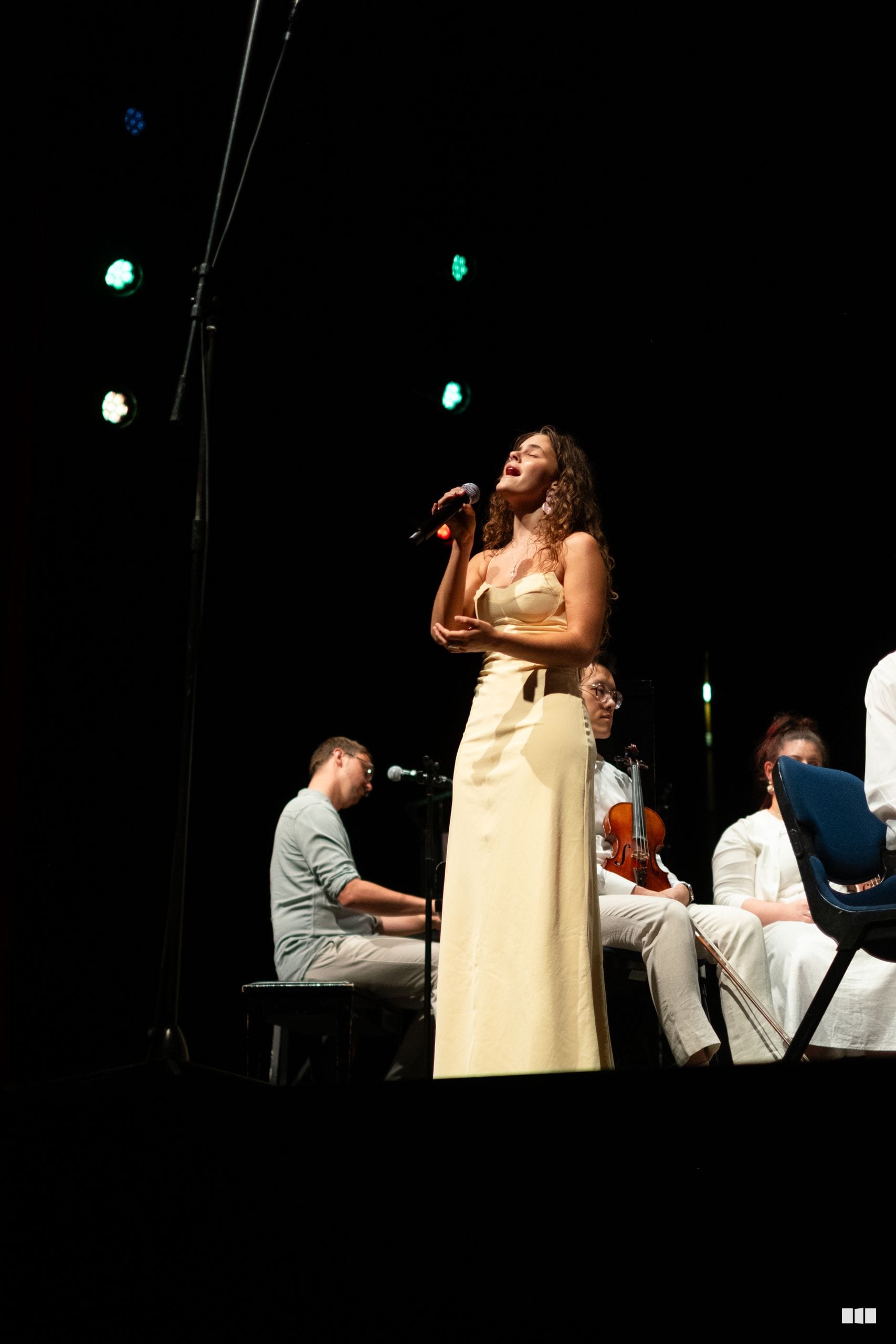 A female soloist in a cream dress performs with a pianist and string musicians on stage.