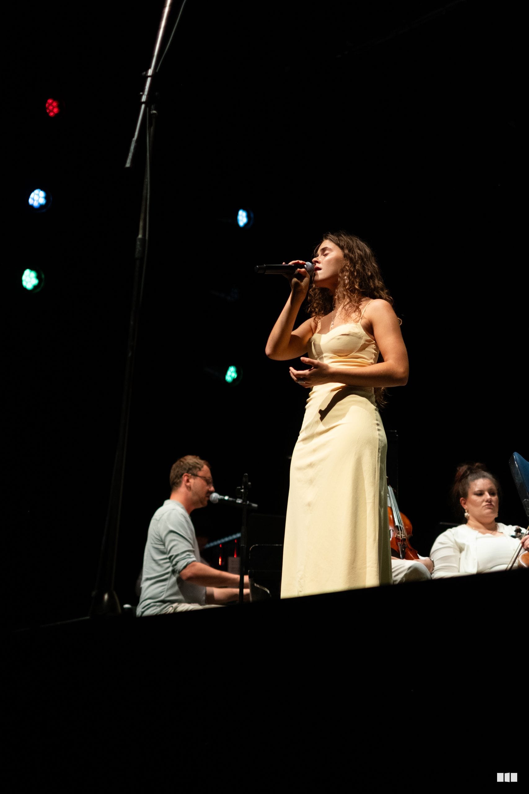 A female soloist in a cream-colored dress performs at the microphone while accompanying musicians play piano and strings in the background.