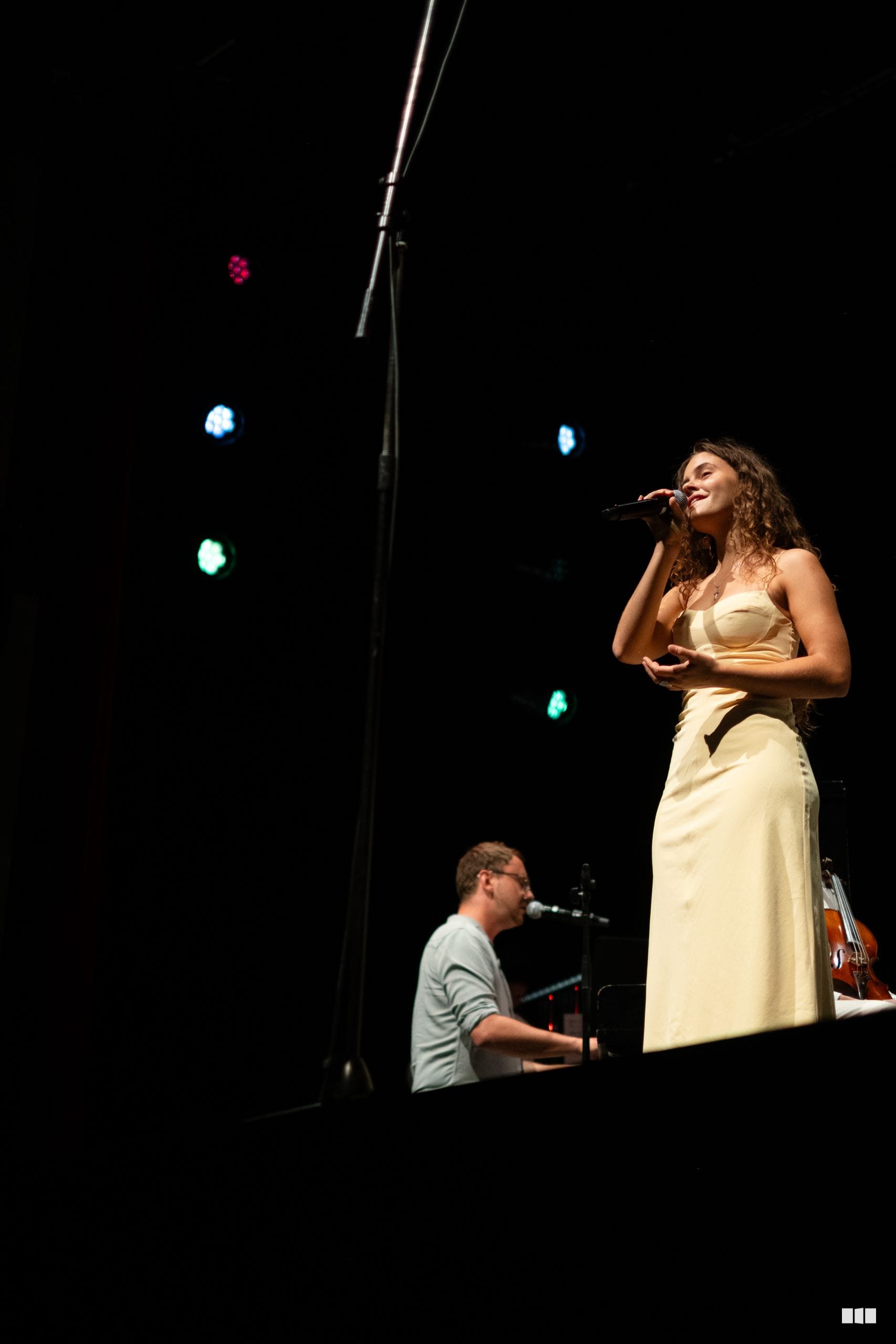 A female soloist in a cream-colored dress performs at the microphone while a pianist accompanies her on stage.