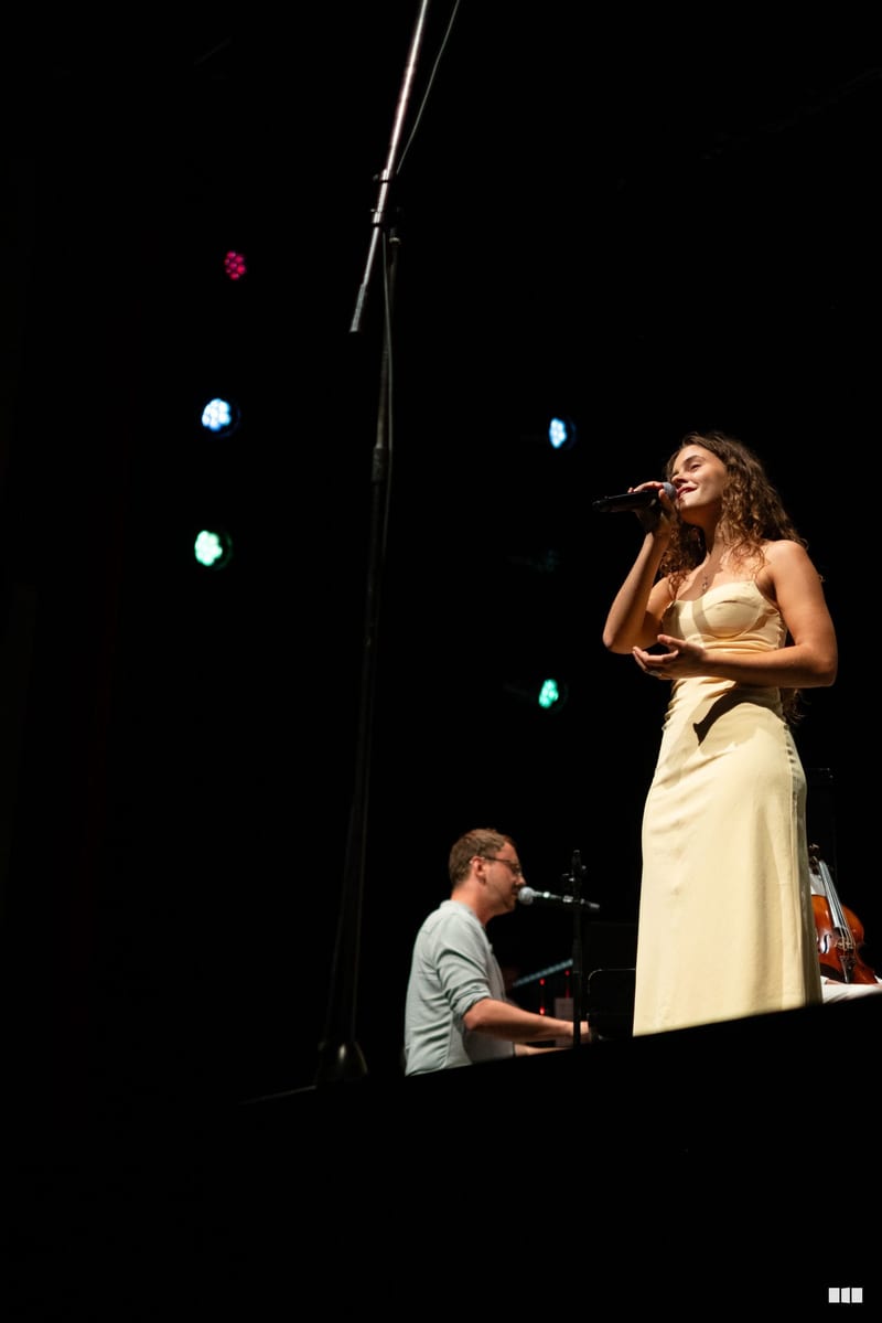 A female soloist in a cream-colored dress performs at the microphone while a pianist accompanies her on stage.