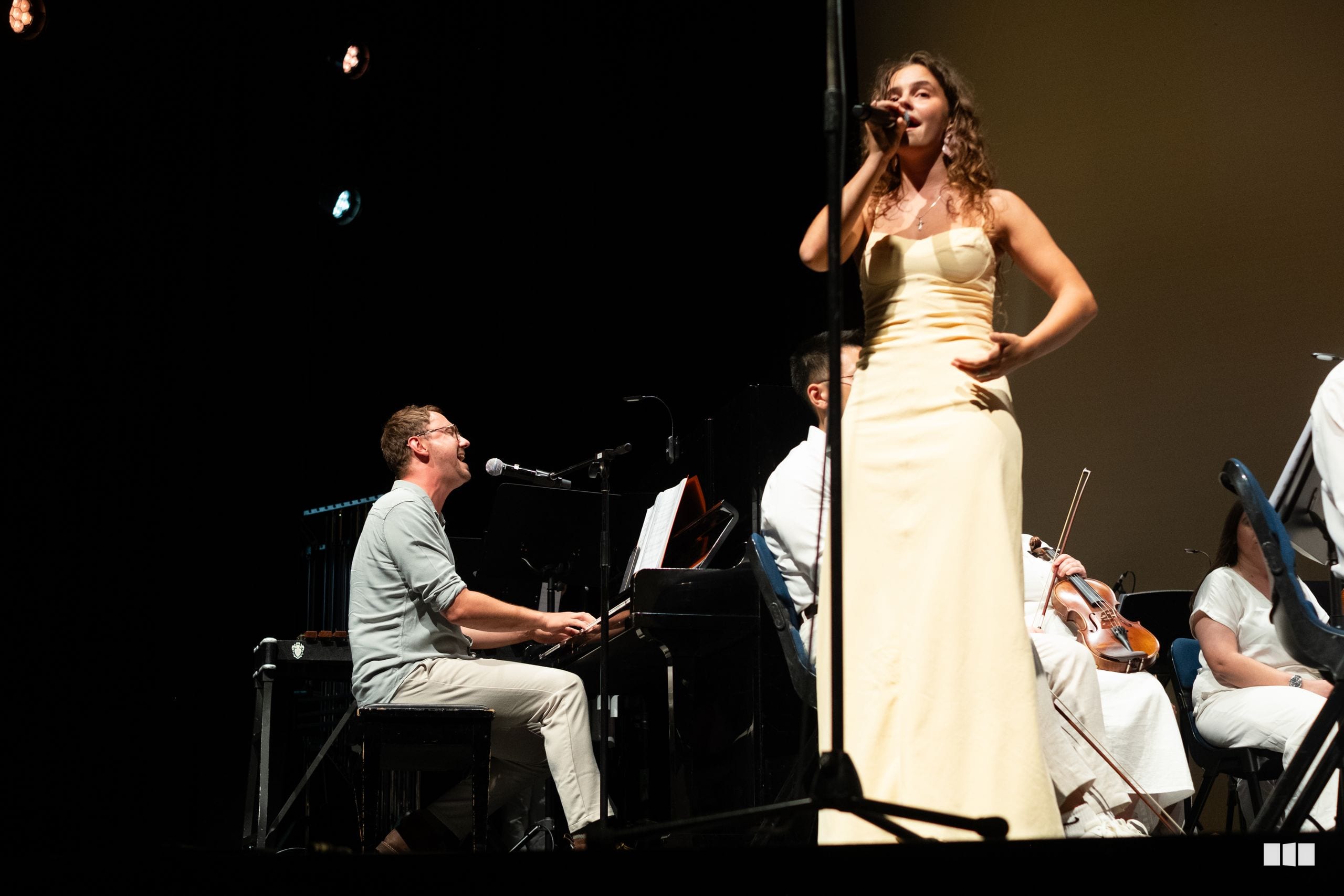 A soprano soloist in a cream gown performs with piano accompaniment and string players during an orchestral concert.