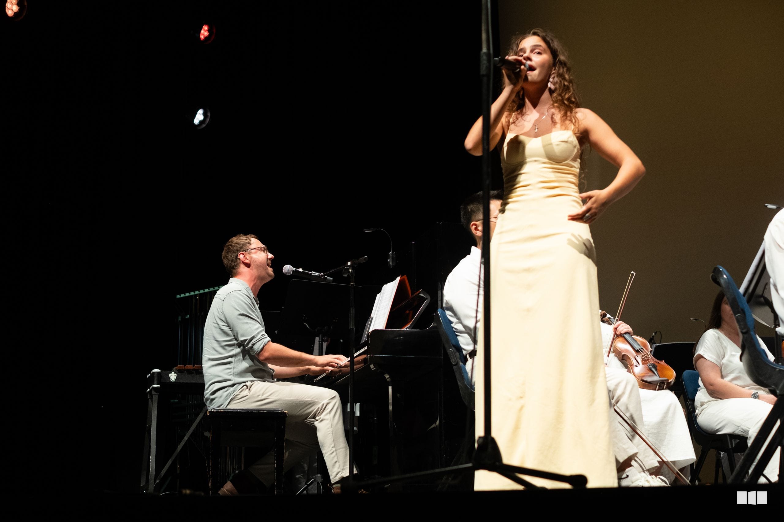 A female soloist in a cream gown performs with a pianist and string musicians on stage.