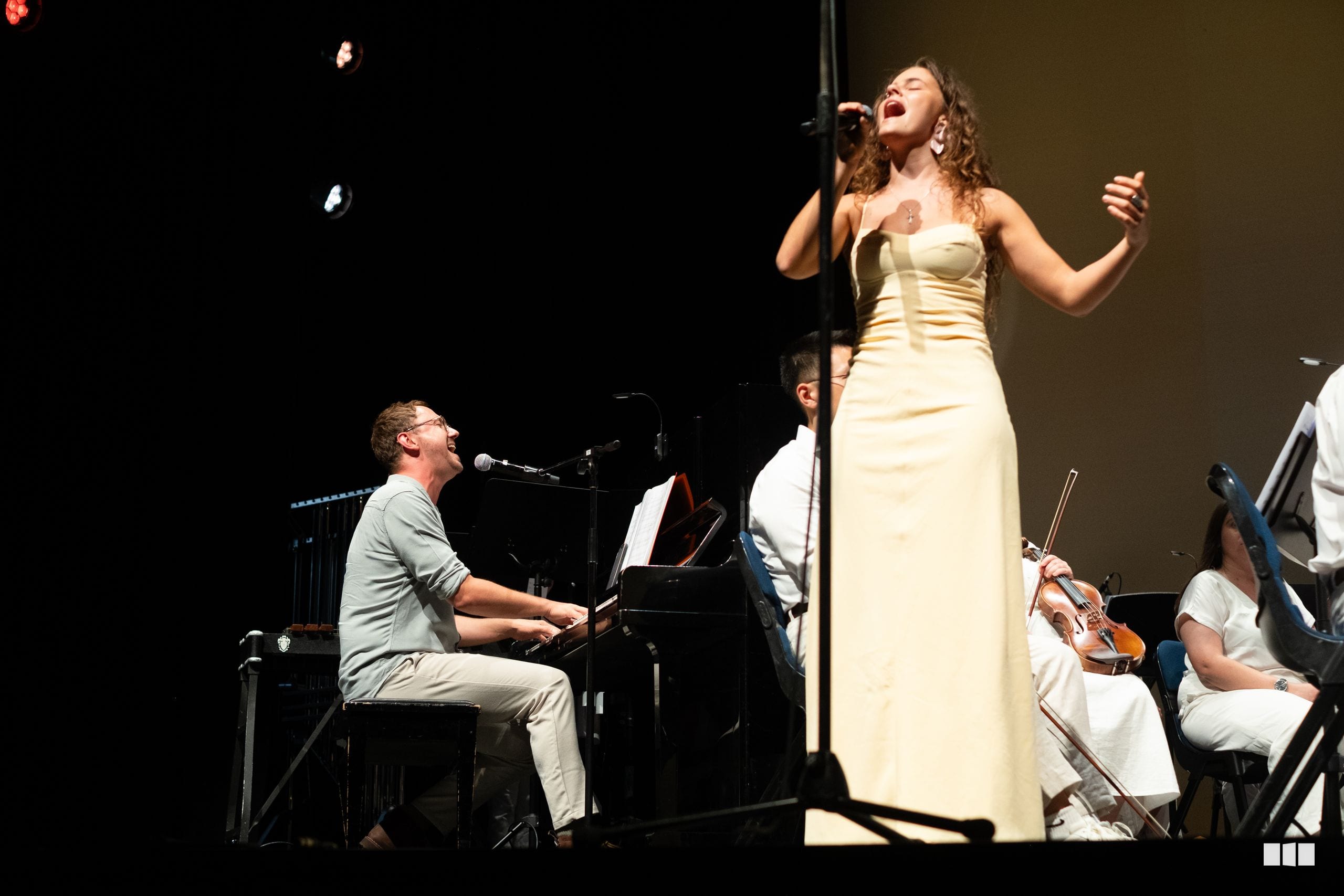 A soprano soloist in an elegant cream gown performs with piano accompaniment and string musicians on stage.