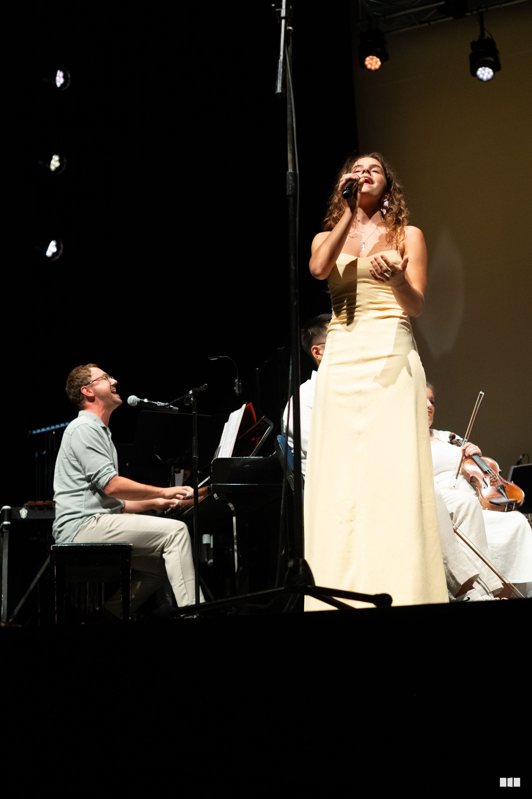 A female soloist in a cream-colored gown performs with a pianist and violinist accompanying her on stage.