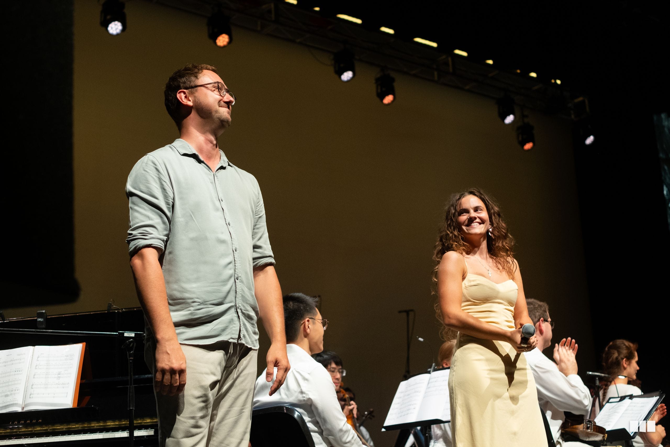 Two soloists take a bow on stage during an orchestral performance, with musicians and sheet music visible in the background.