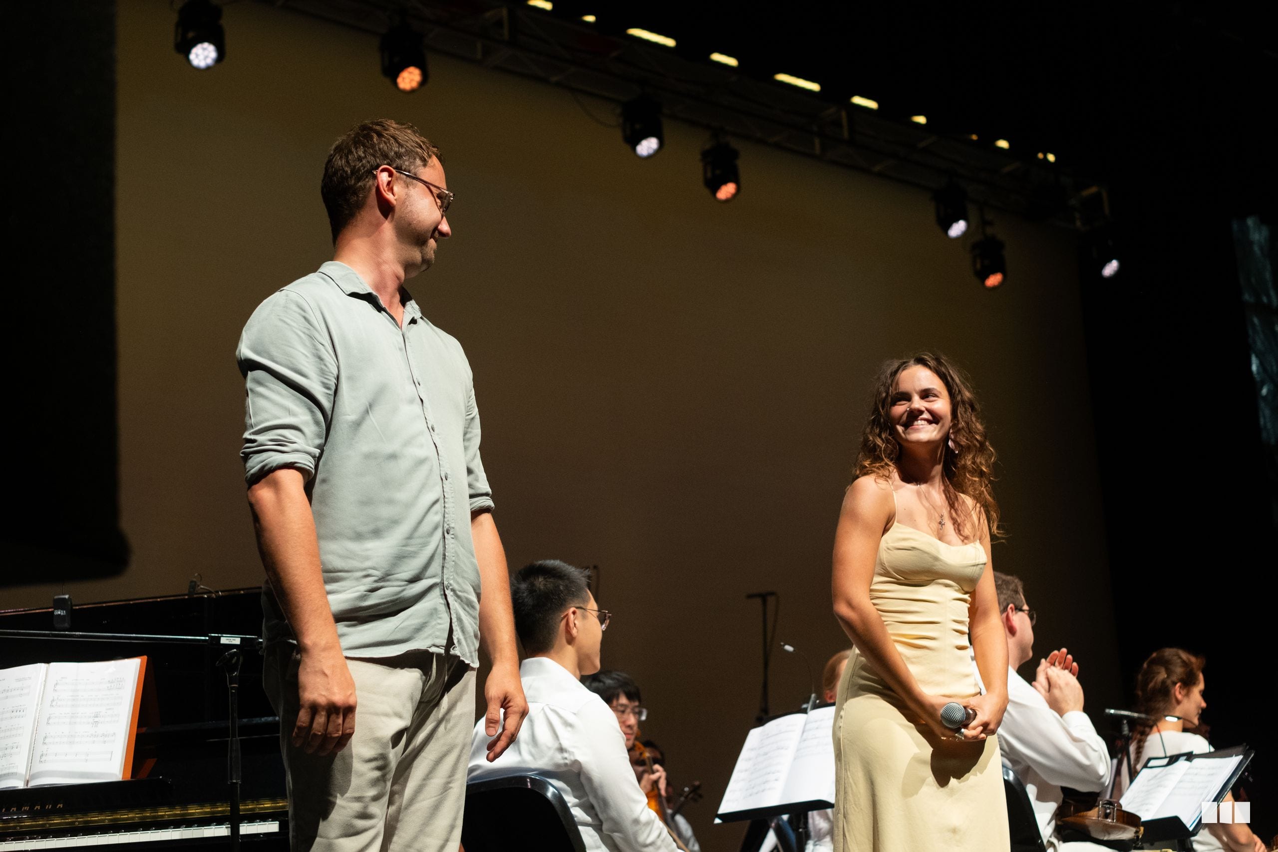 A male conductor and female soloist stand together on stage during a performance with the orchestra seated behind them.