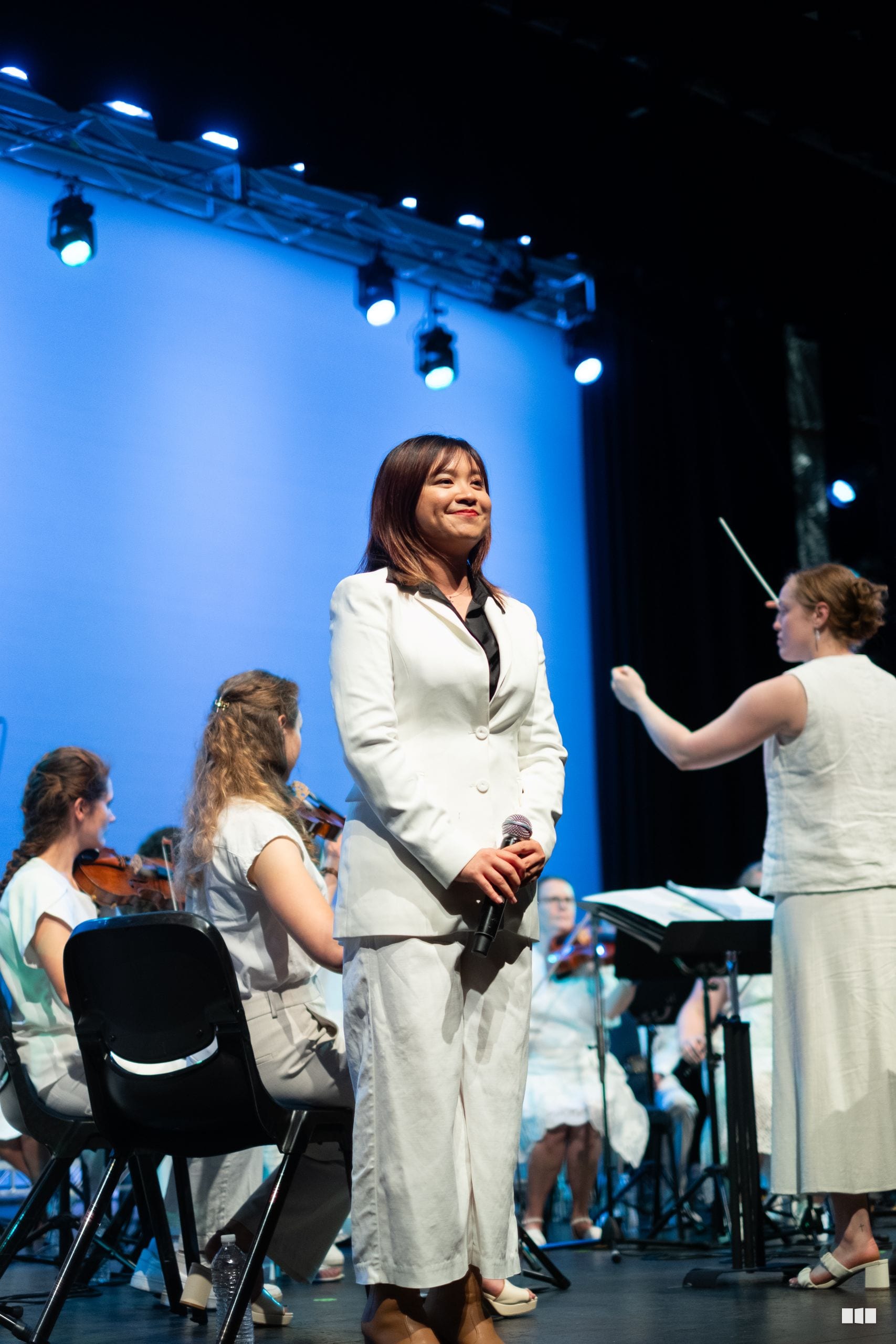 A woman in a white blazer holds a microphone on stage while young musicians with instruments sit nearby under blue stage lighting.