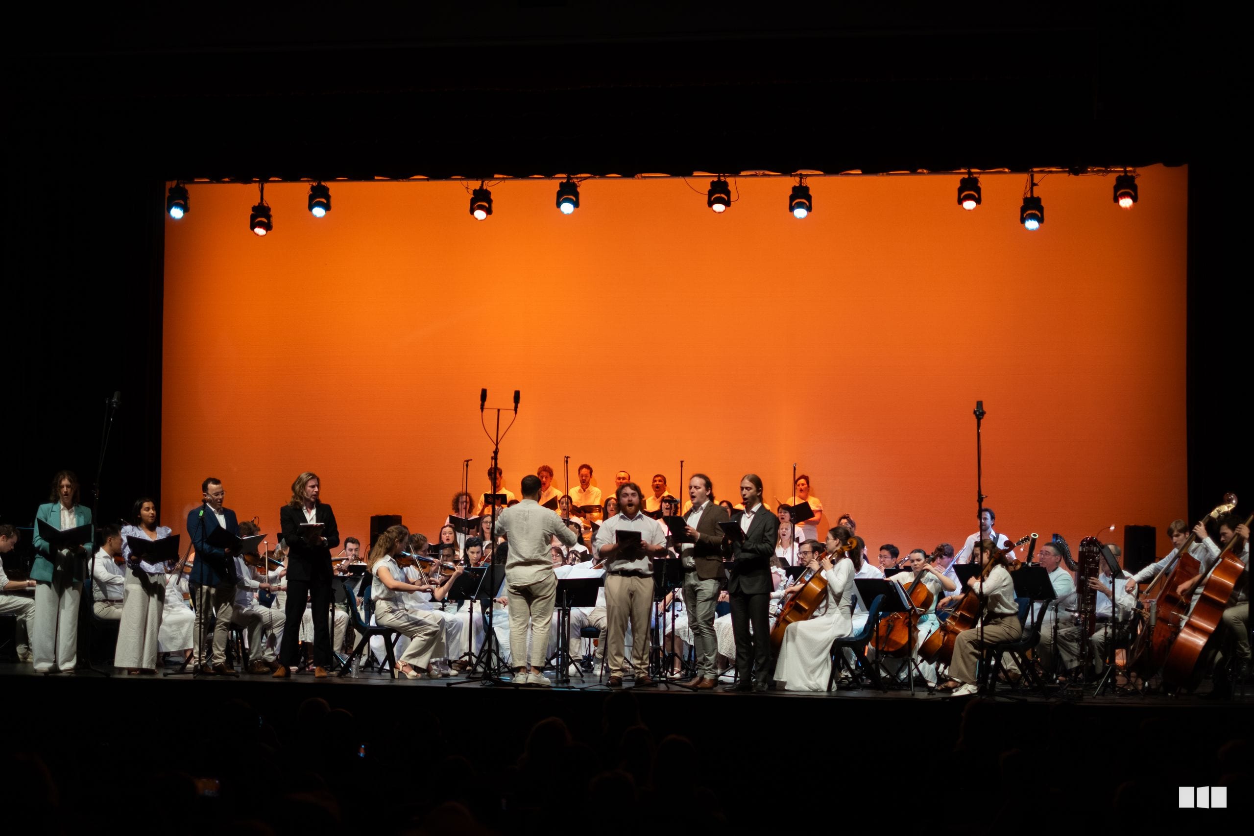 A conductor leads the Australian Christian Arts orchestra and choir in a dynamic performance on stage beneath vibrant orange lighting.