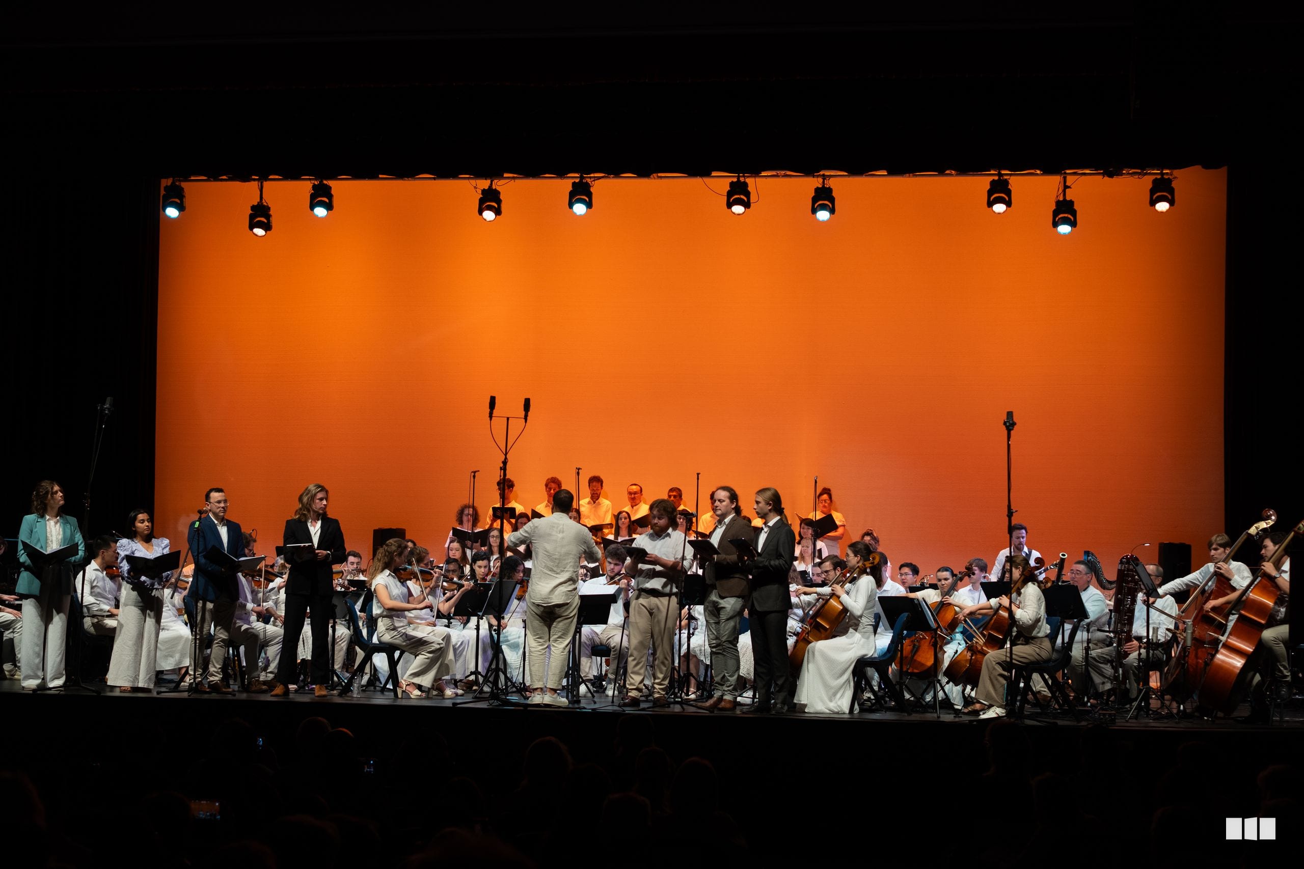 A conductor leads a full orchestra with choir members in the background against a vibrant orange backdrop during a performance.