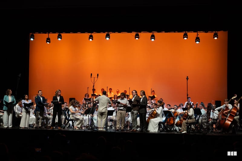 A conductor leads a full orchestra with choir members in the background against a vibrant orange backdrop during a performance.