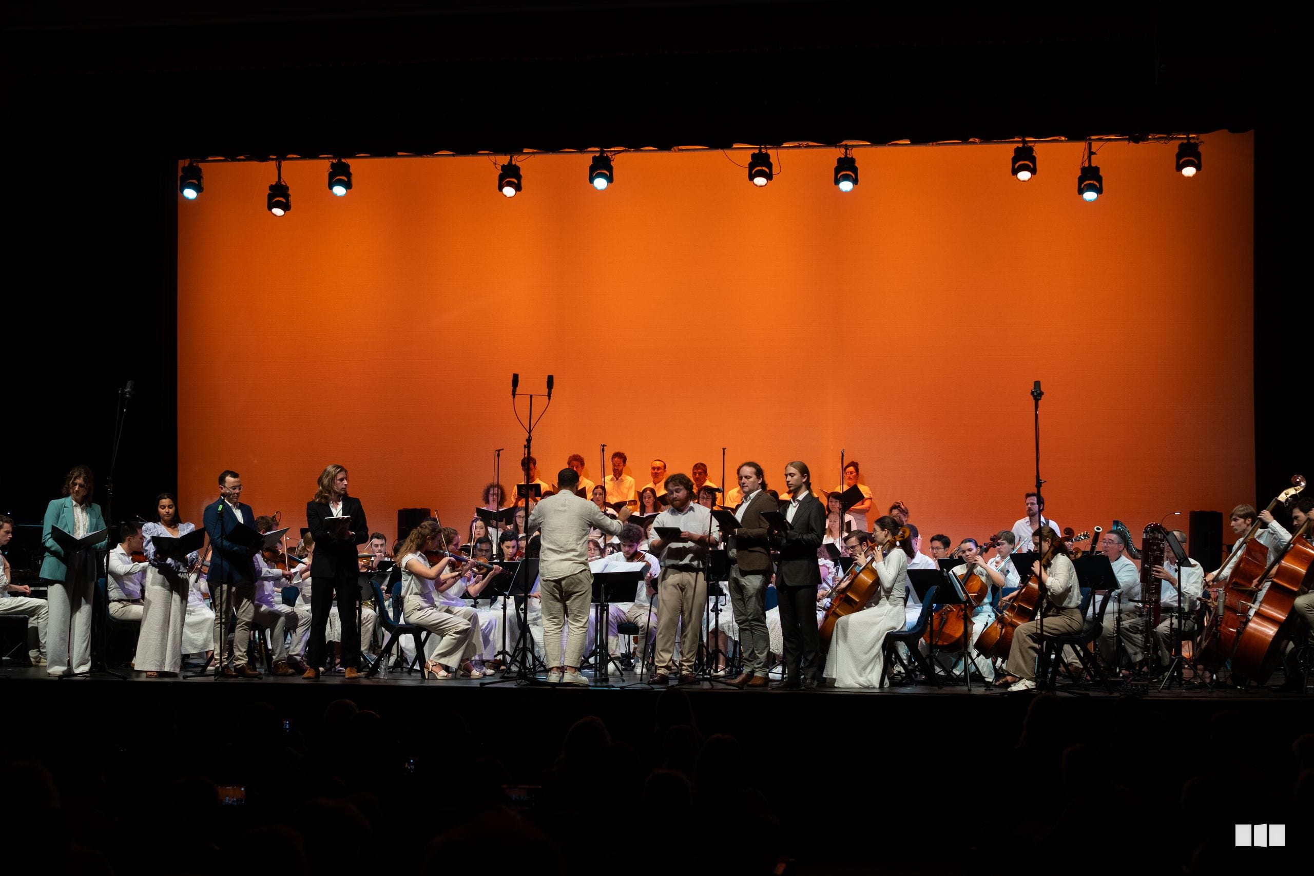 A conductor leads a full orchestra and choir during a performance on stage with dramatic orange backdrop lighting.