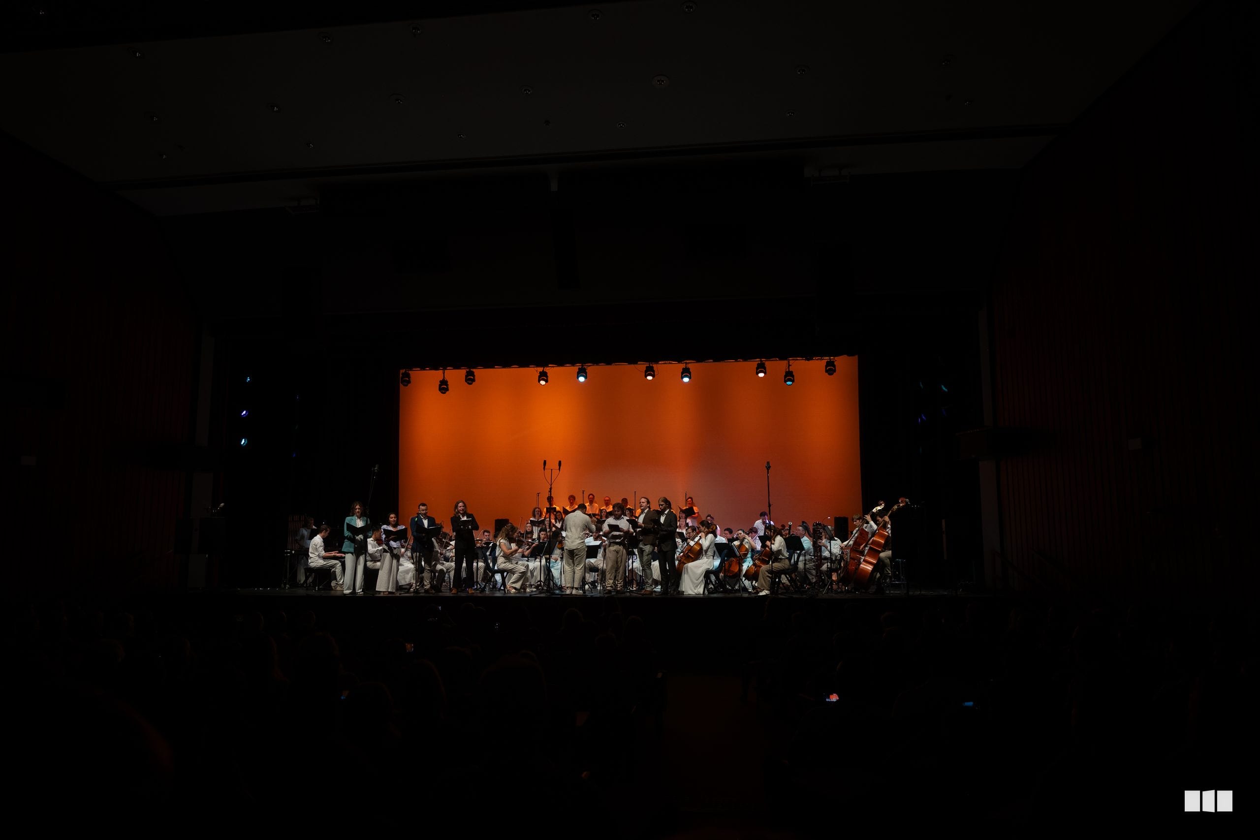 A full orchestra and choir perform together on stage under warm orange lighting during a concert presentation.
