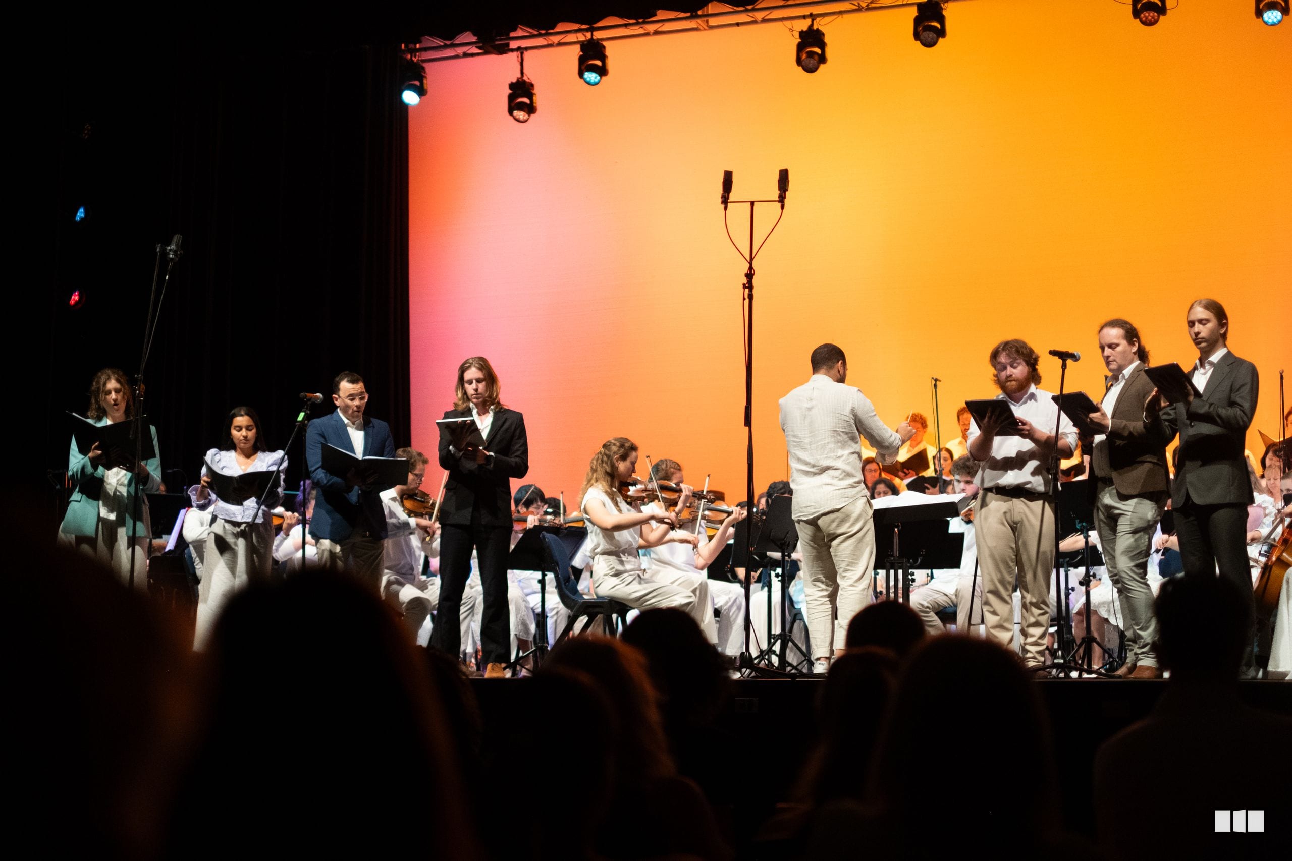 Orchestra and soloists take a bow during a concert performance with warm orange stage lighting.