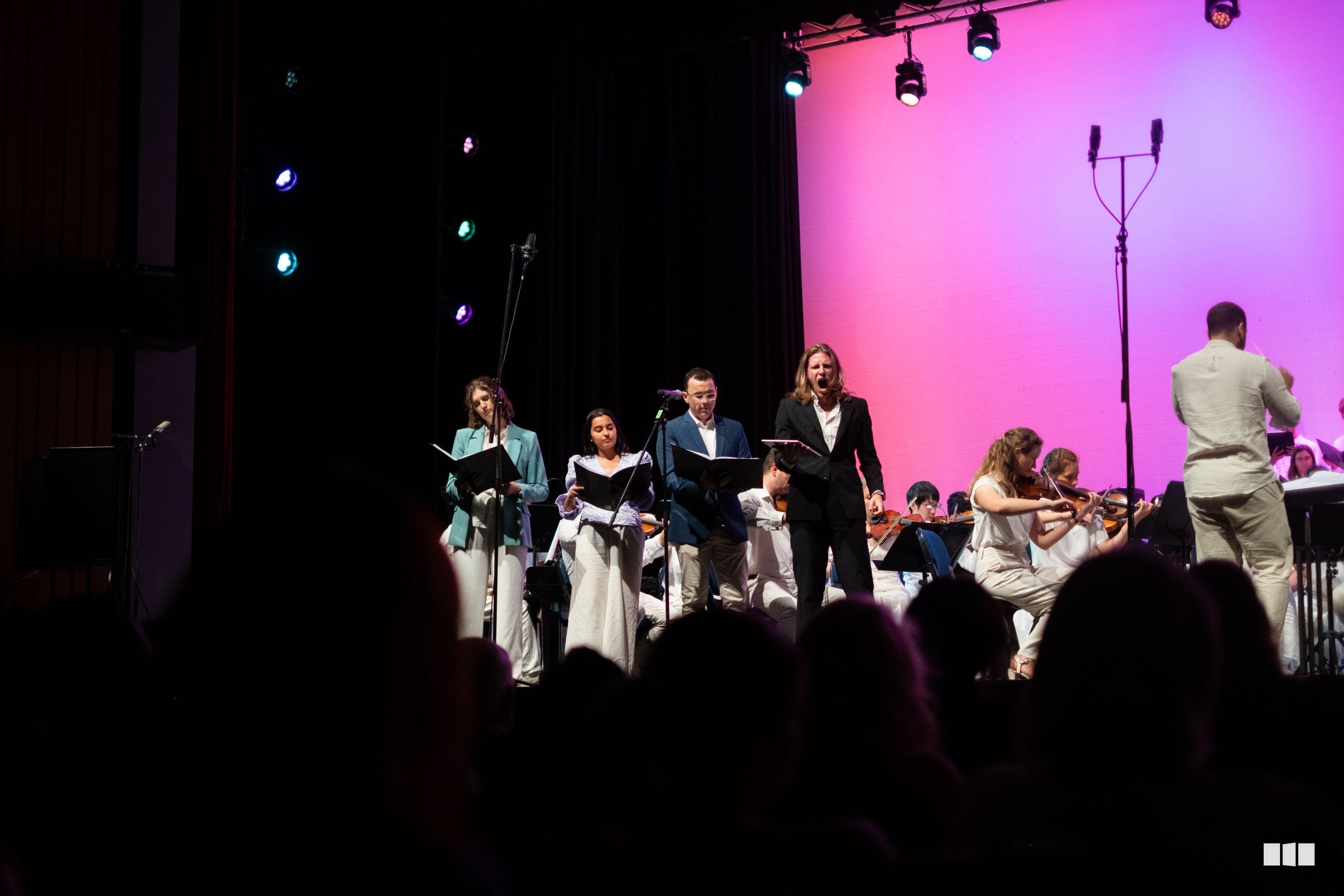 Young musicians and singers perform together on stage with dramatic pink and purple lighting during an orchestral and choral performance.