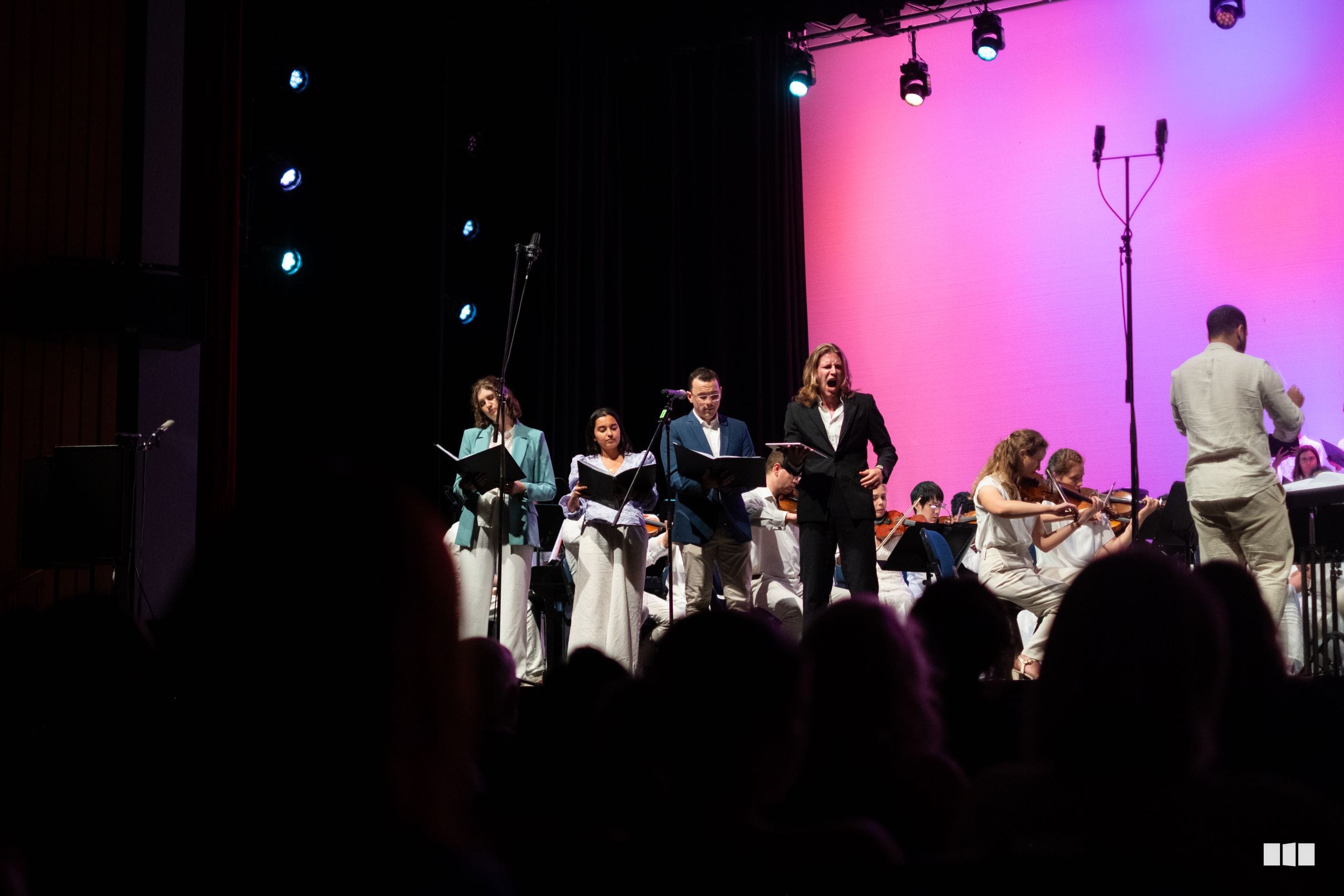 Young musicians perform with adult soloists and singers on stage during an orchestral and choral performance under pink and purple stage lighting.