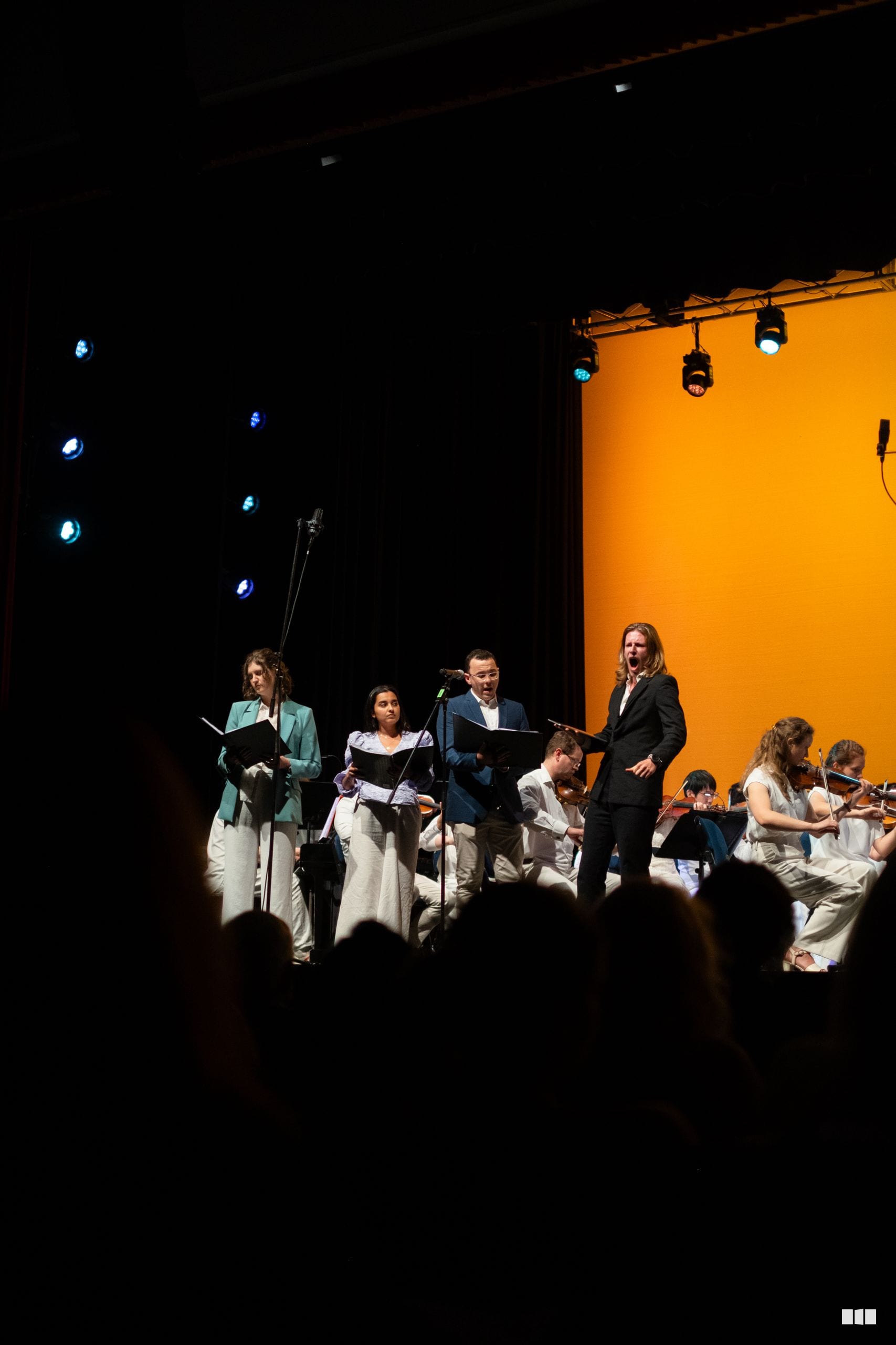 Soloists and orchestra members perform together on stage with sheet music under stage lighting.