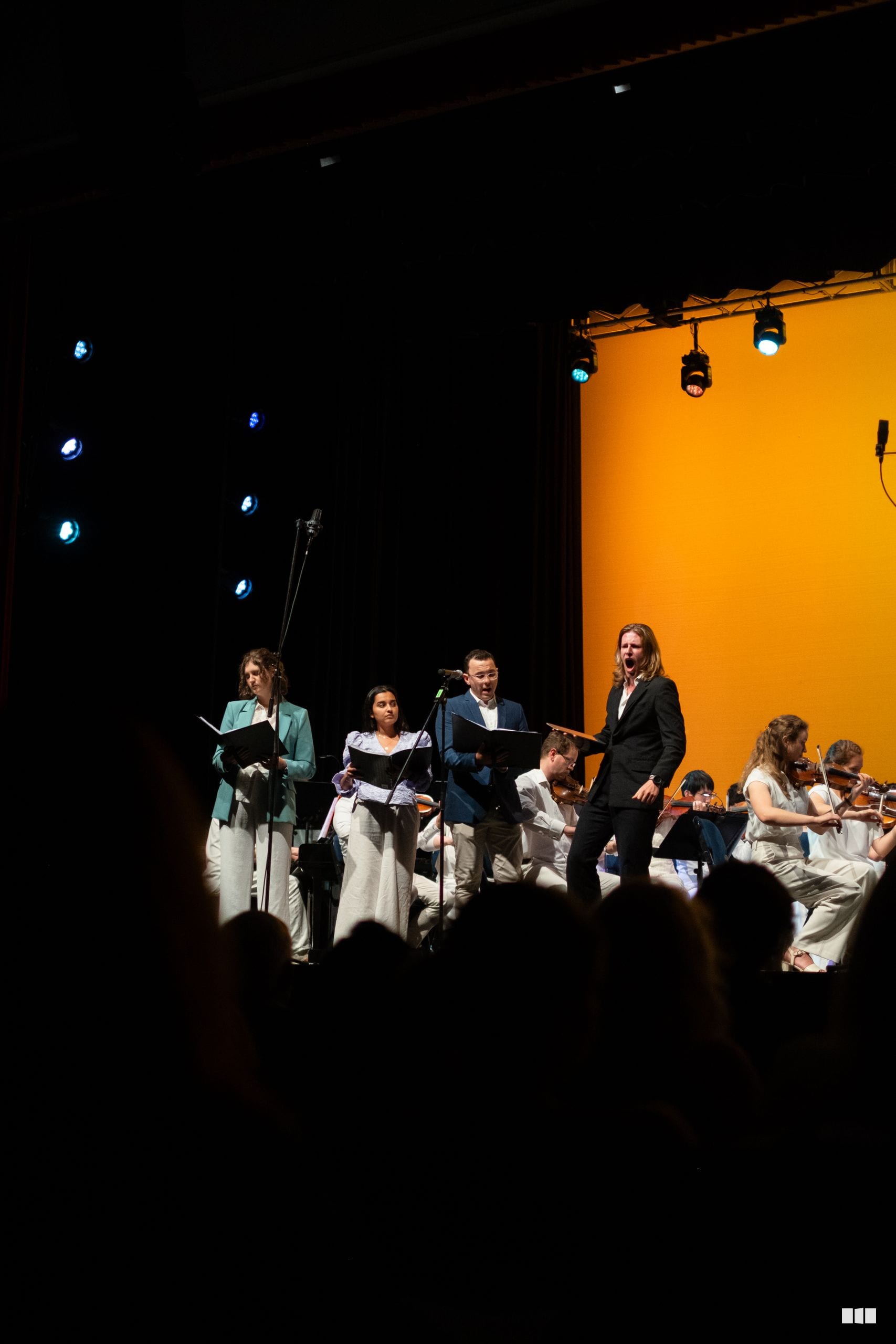 Choral singers in formal attire perform on stage with orchestra members visible on the right side, accompanied by professional stage lighting and an orange backdrop.