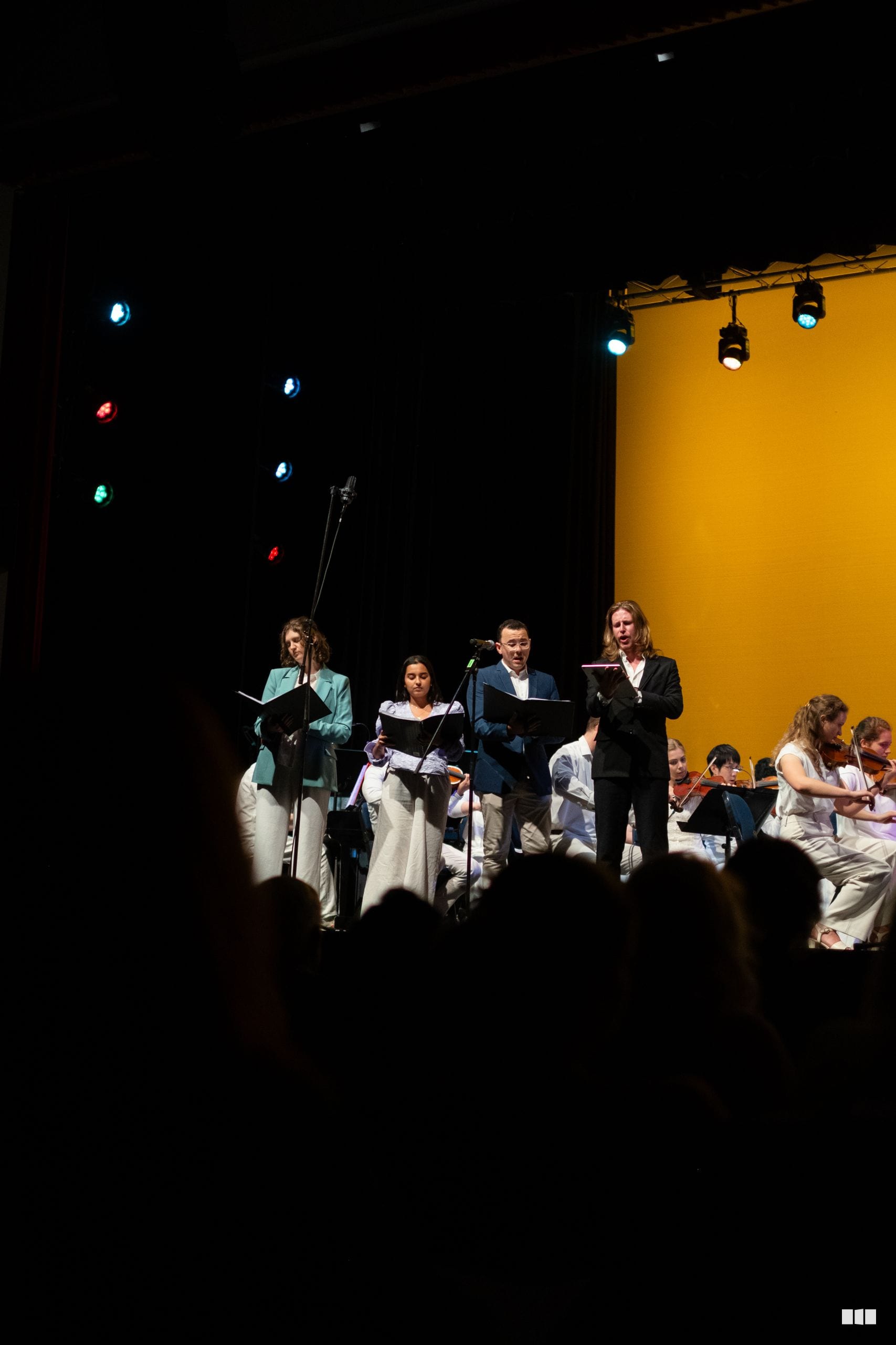 Soloists and orchestra members perform together on stage with sheet music under dramatic stage lighting.