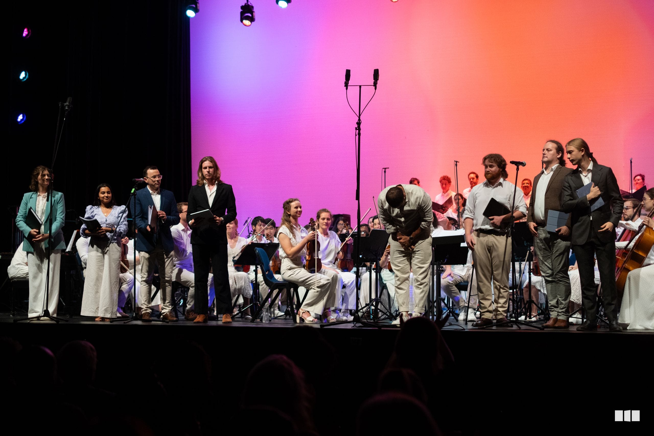 Orchestra and choir members take a bow together on stage under vibrant pink and orange lighting after a performance.