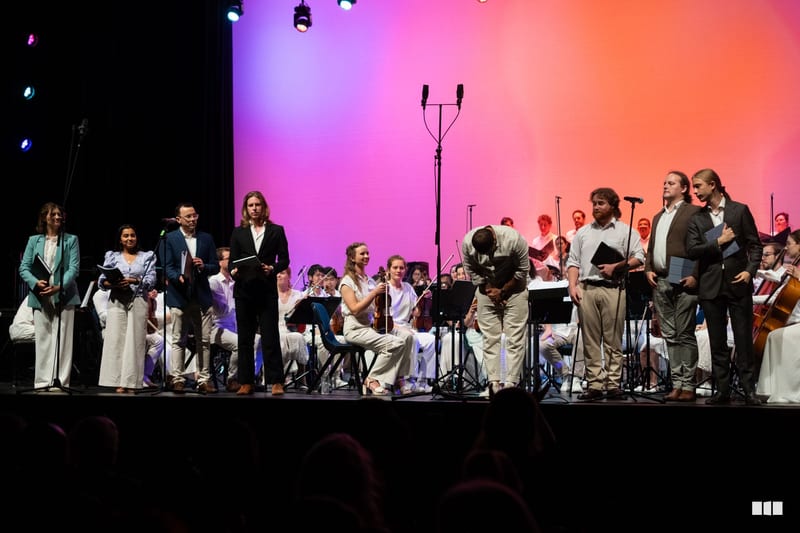 Orchestra and choir members take a bow together on stage under vibrant pink and orange lighting after a performance.