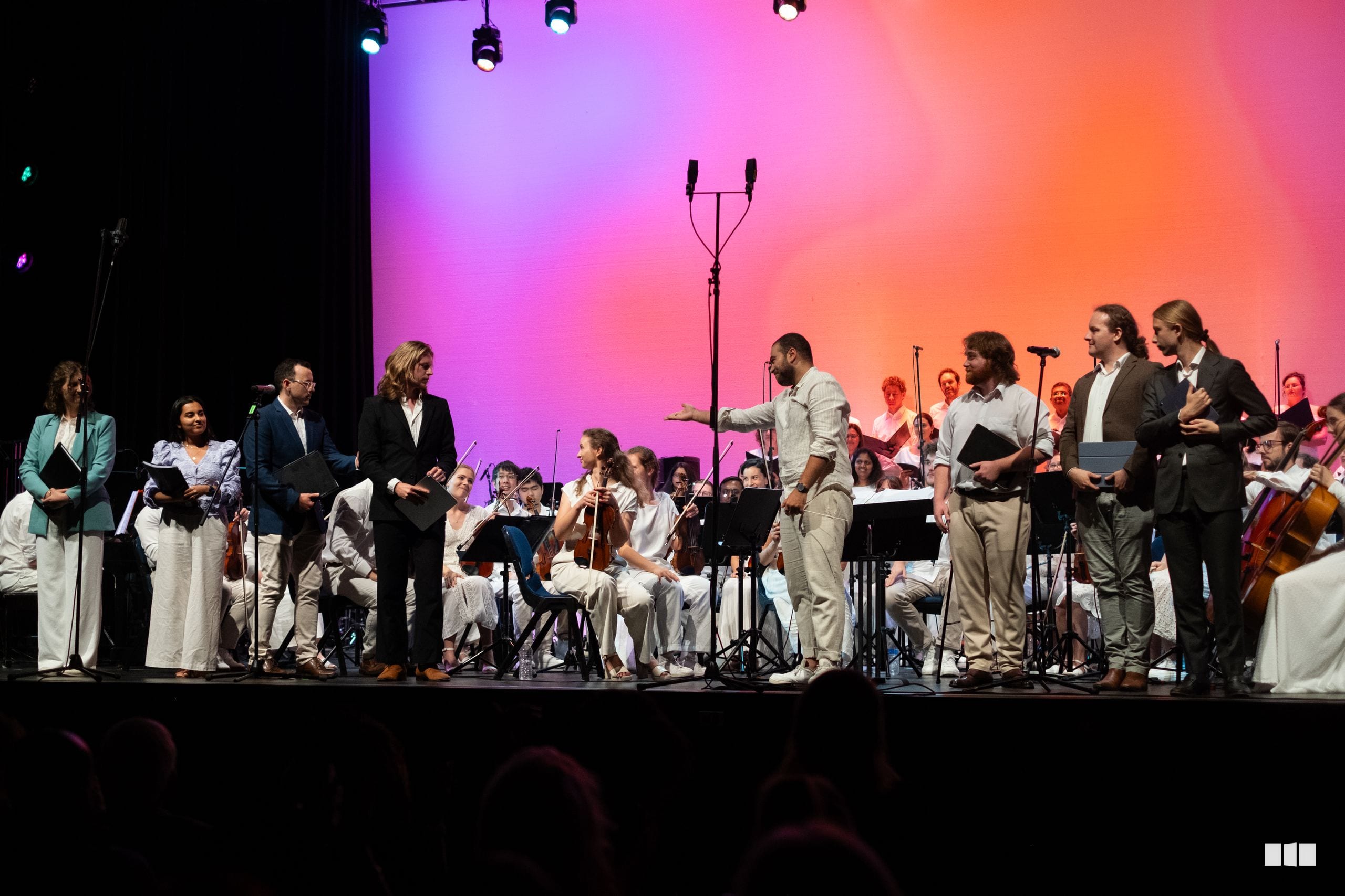 Orchestra and choir members take their bows on stage during a performance, with musicians and singers arranged across the platform under vibrant pink and purple stage lighting.