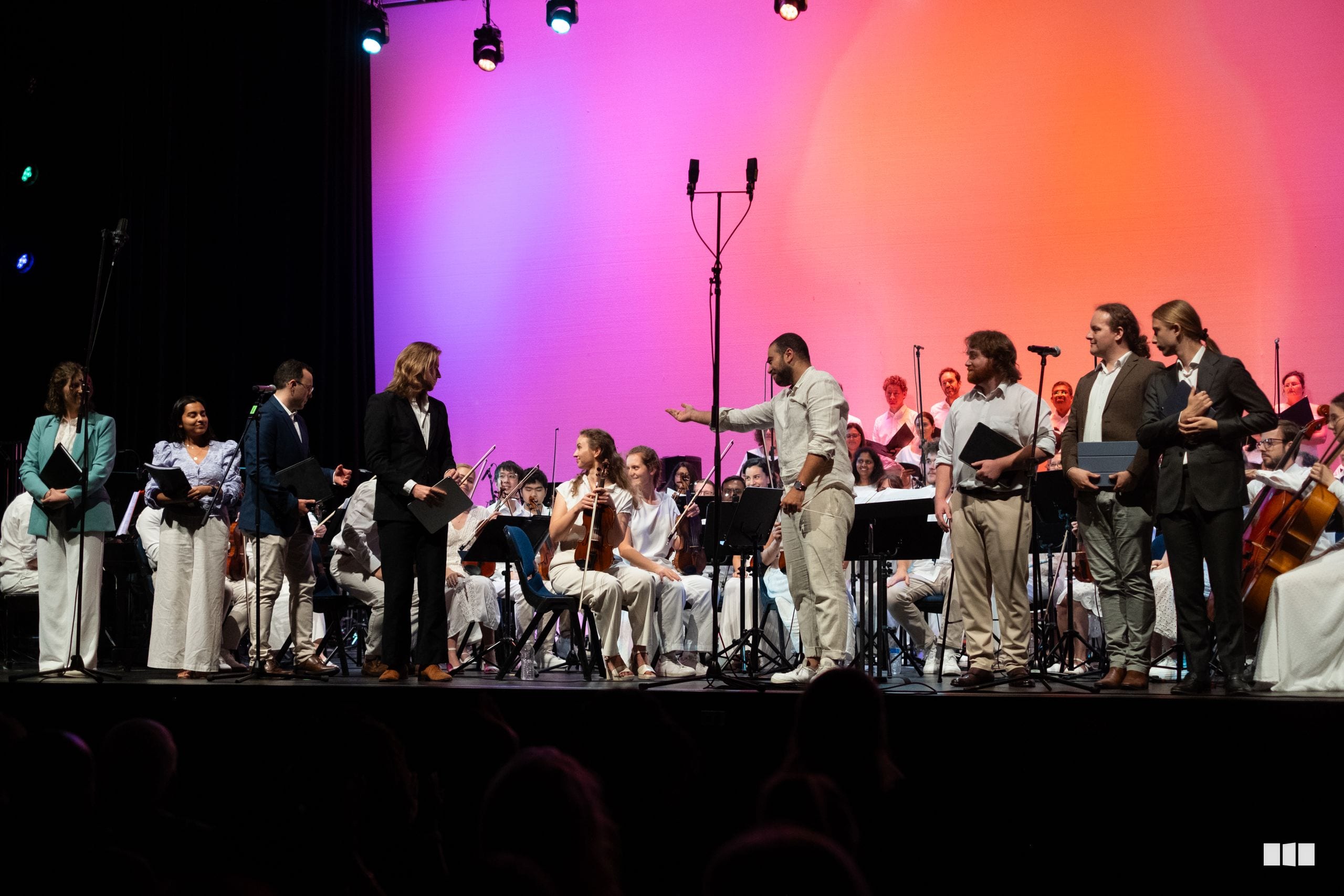 A diverse group of orchestral musicians and singers perform together on stage under vibrant pink and purple lighting during a concert.
