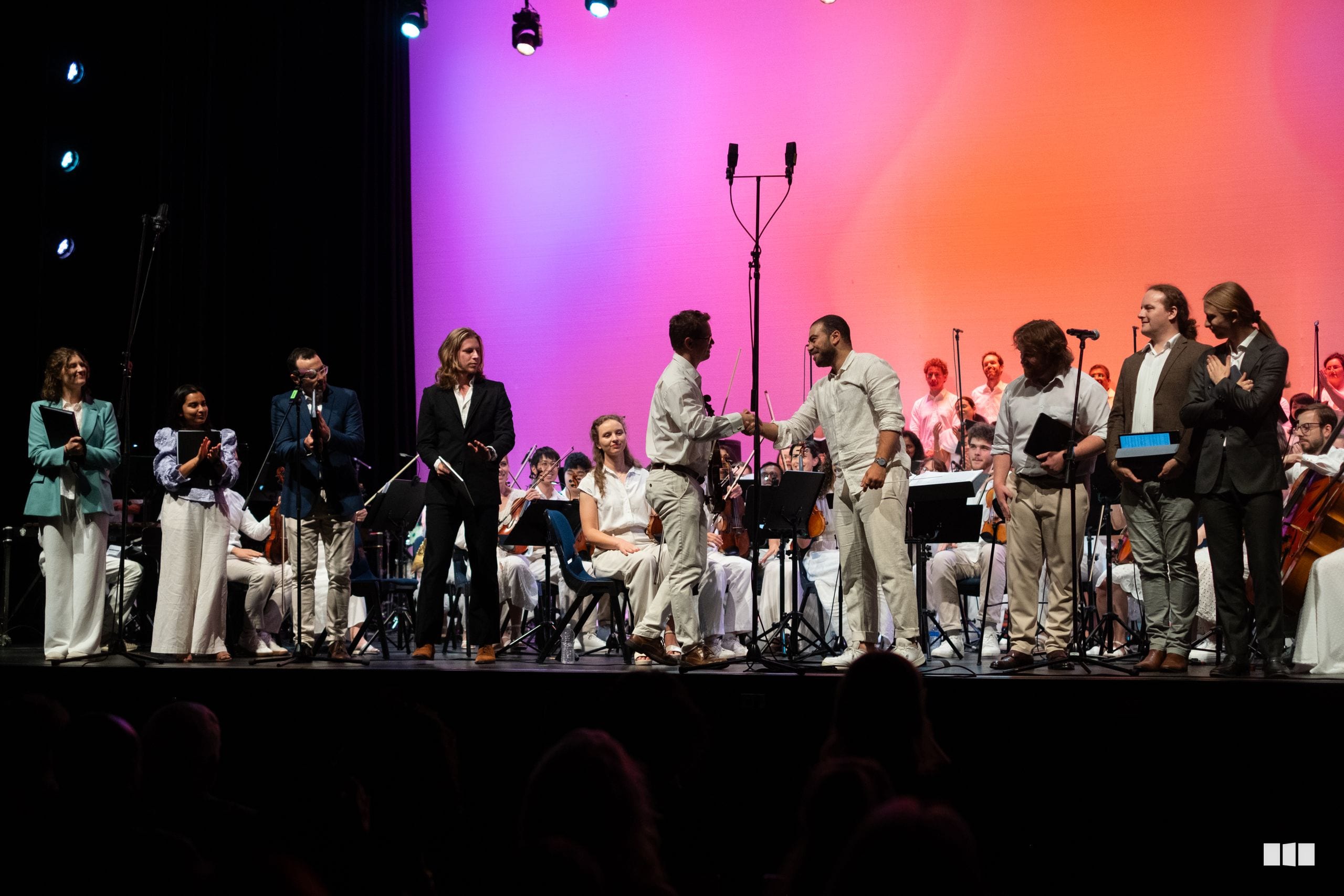 Orchestra and choir members take a bow together on stage under vibrant purple and orange lighting after a performance.