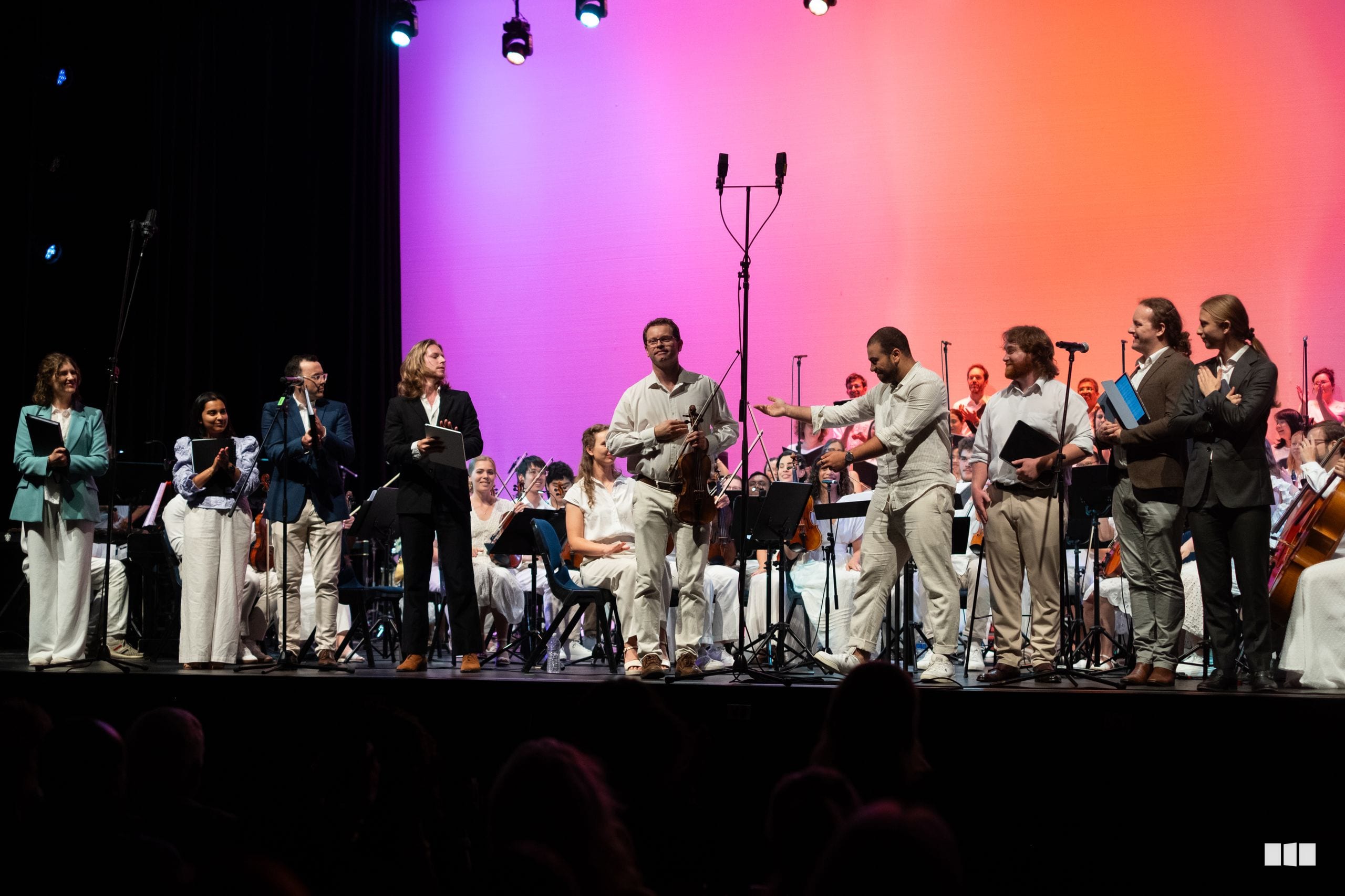 Orchestra and choir members stand together on stage during a performance with vibrant pink and orange lighting.