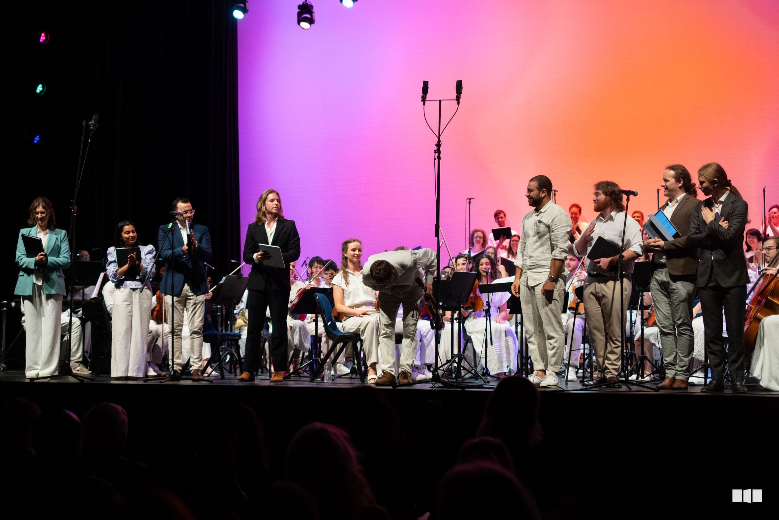 A mixed ensemble of singers and orchestra members take a bow together on stage under vibrant pink and purple lighting.