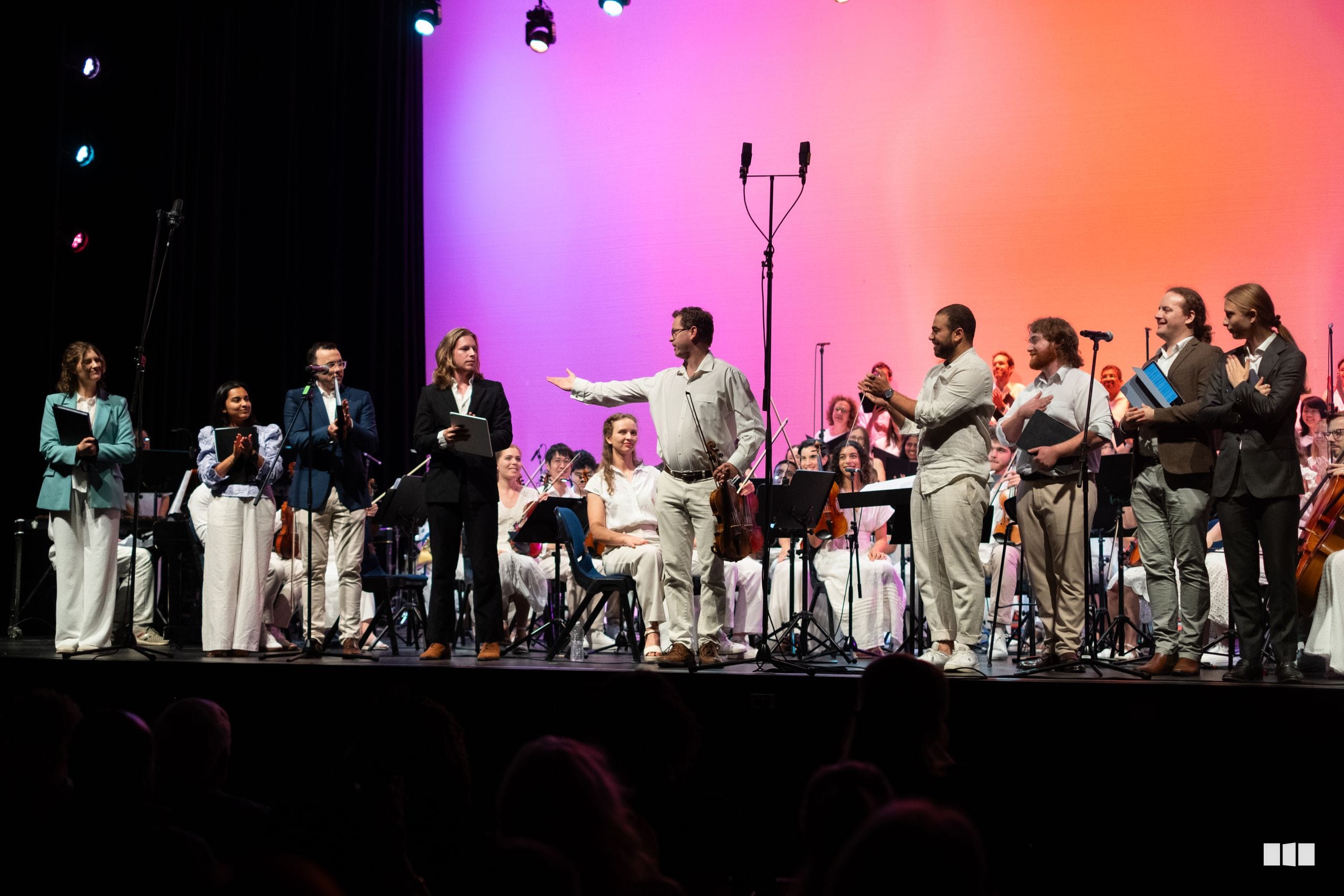 A conductor leads an orchestra and choir in an open rehearsal or concert performance on stage with vibrant pink and orange lighting.