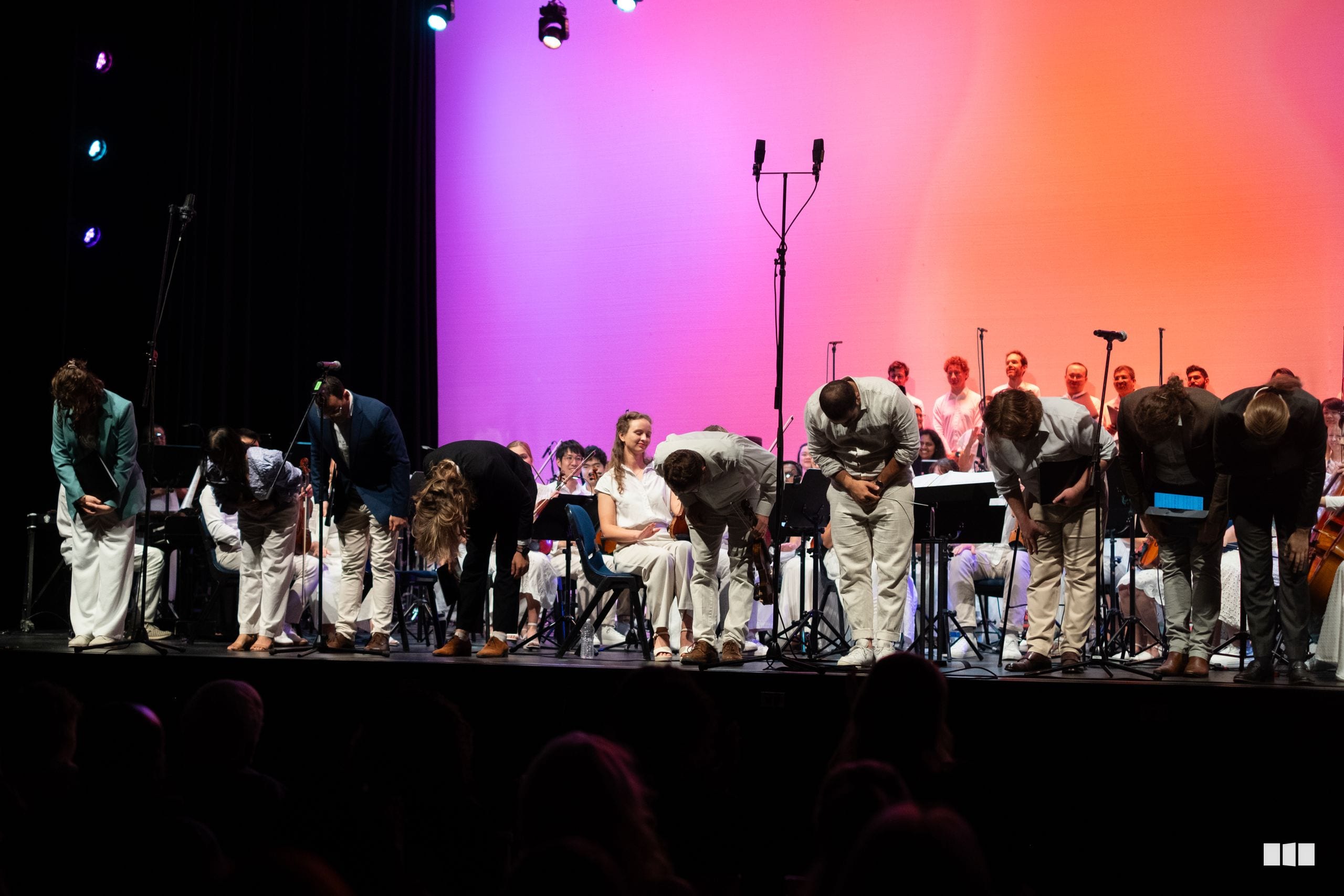 Orchestra and choir members take a bow together on stage under vibrant pink and orange lighting at the conclusion of a performance.