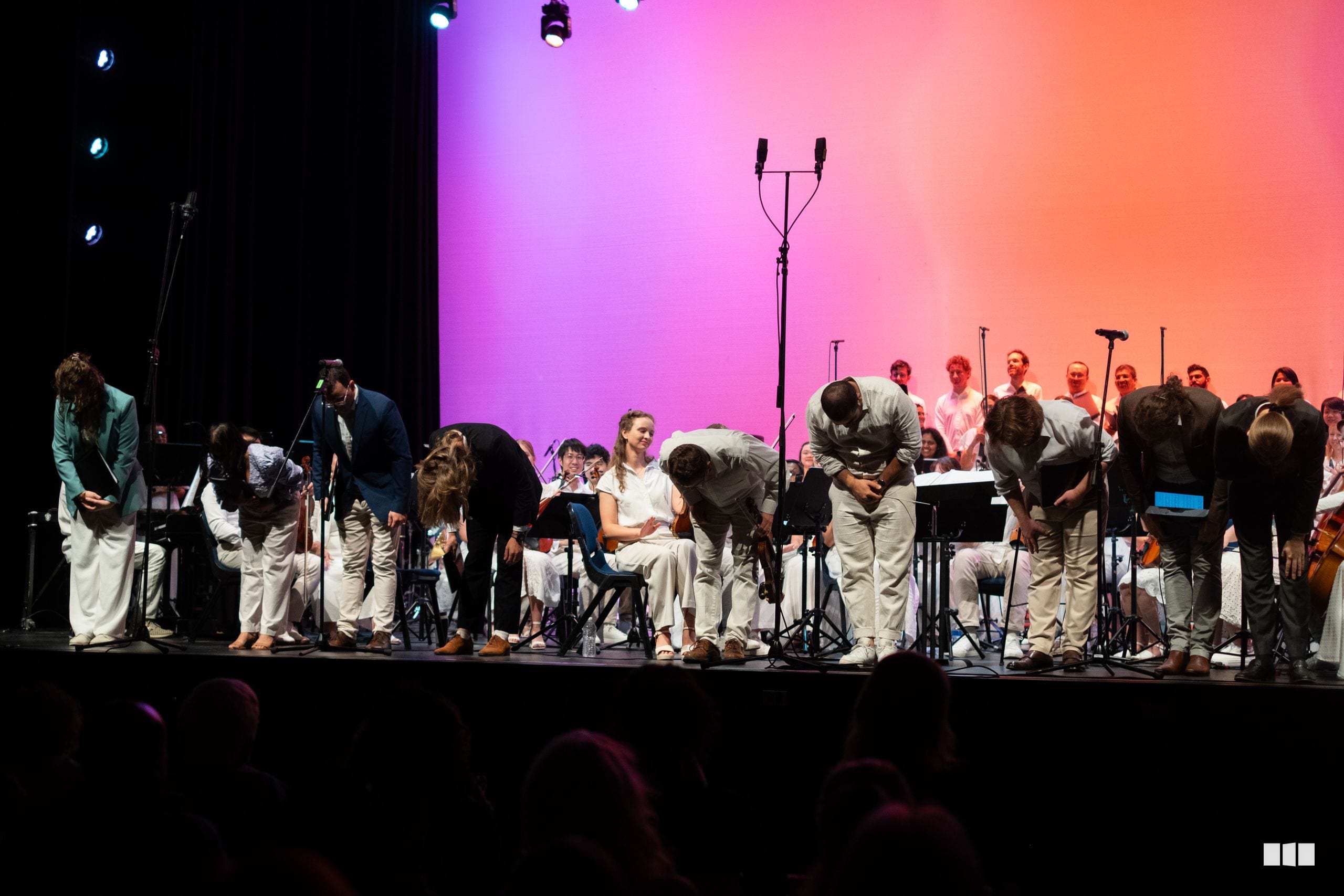 Orchestra and choir members take a bow together on stage under vibrant pink and orange lighting after a performance.