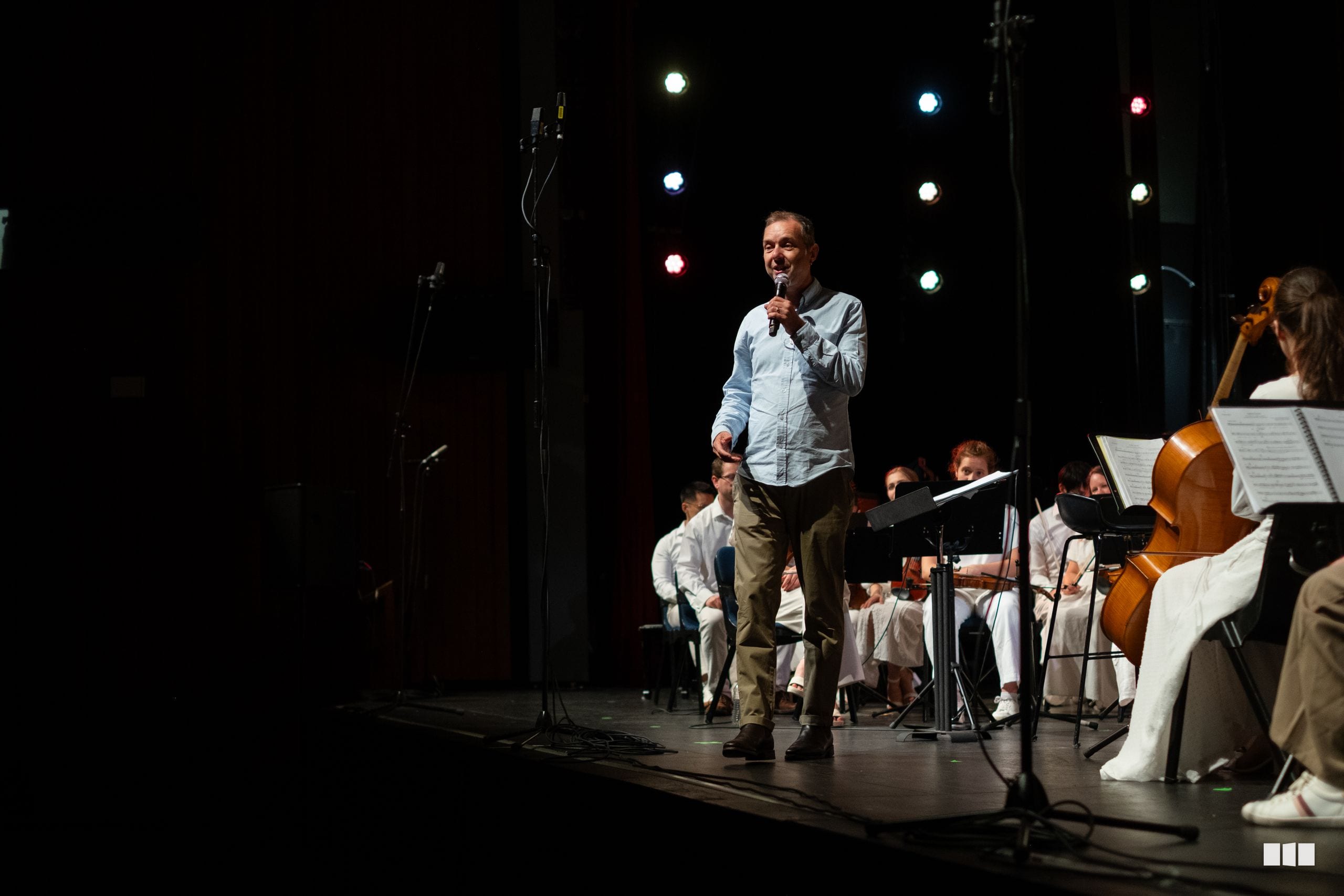 A speaker addresses the audience while orchestra musicians in white shirts perform on stage under colorful stage lighting.