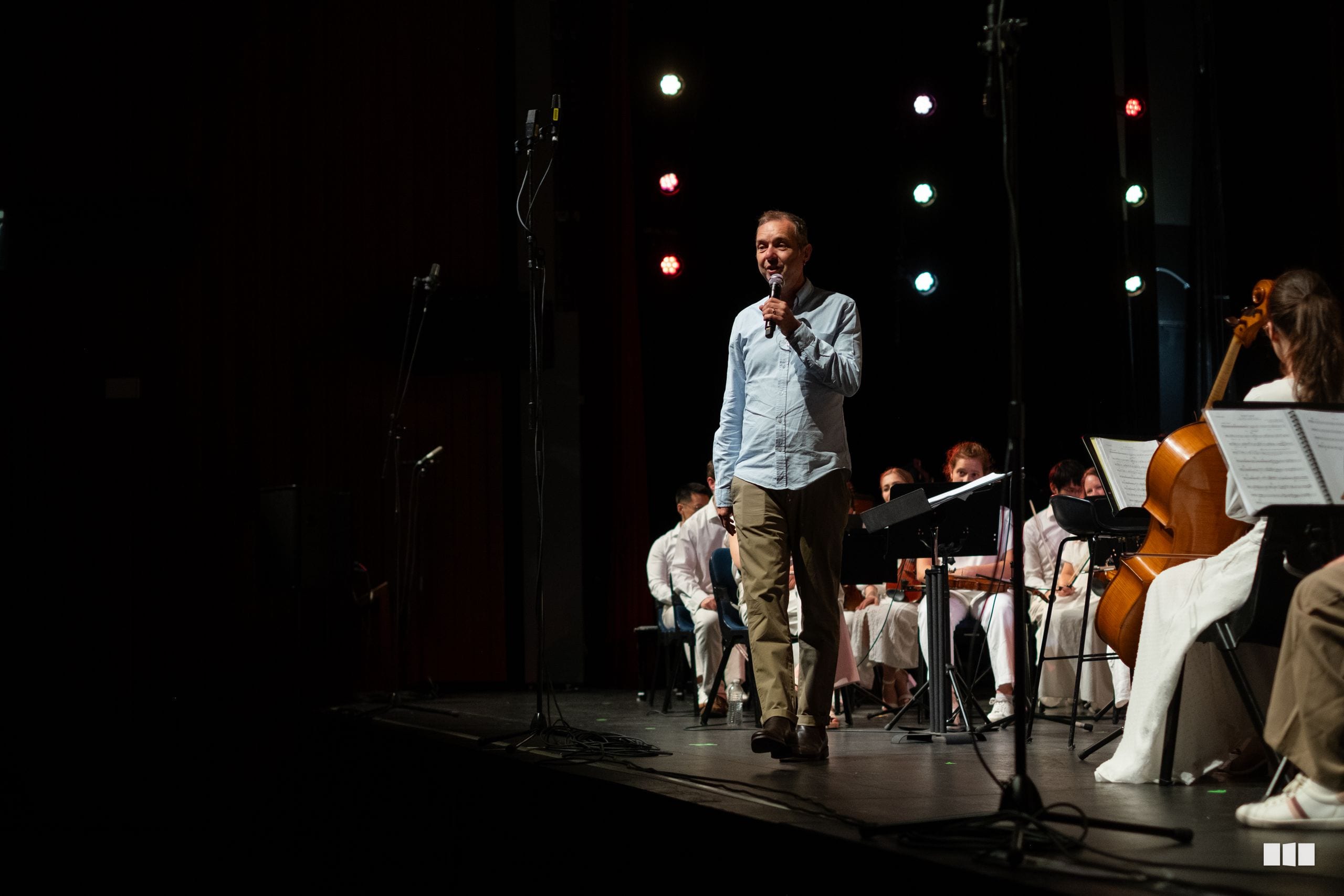 A speaker addresses the audience while an orchestra with string players performs on stage during an evening concert.